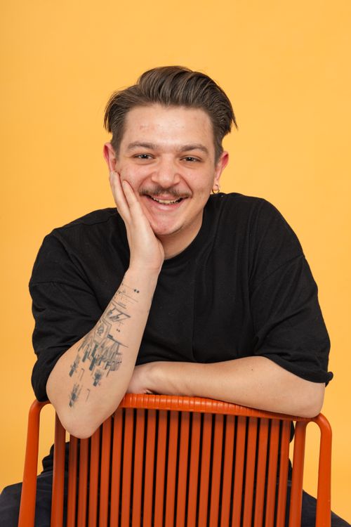 Free A cheerful man with tattoos smiles while seated on an orange chair, set against a bright yellow backdrop. Stock Photo