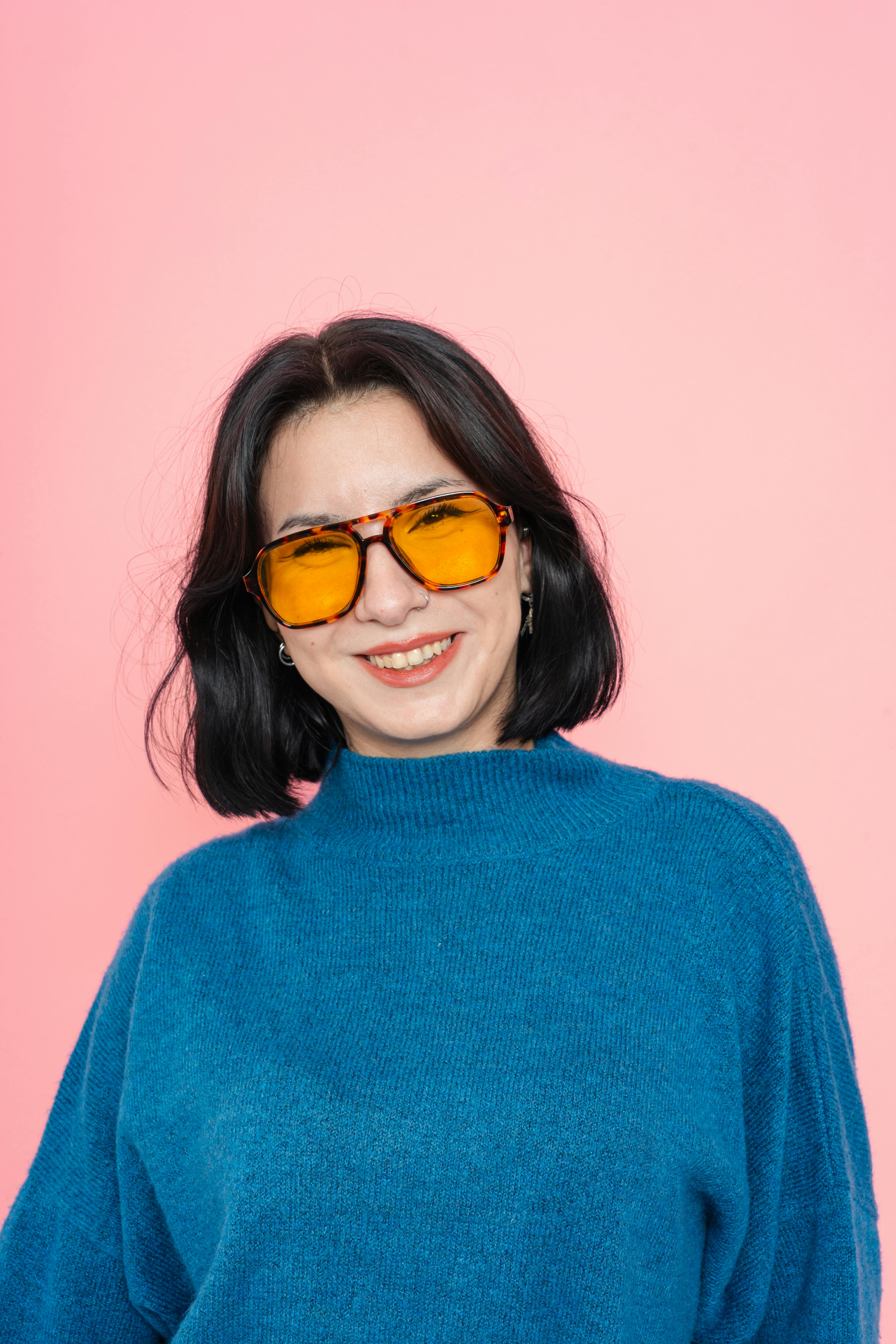 De franc Retrat d'una dona somrient amb ulleres de sol i un jersei blau sobre un fons rosa. Foto d'estoc