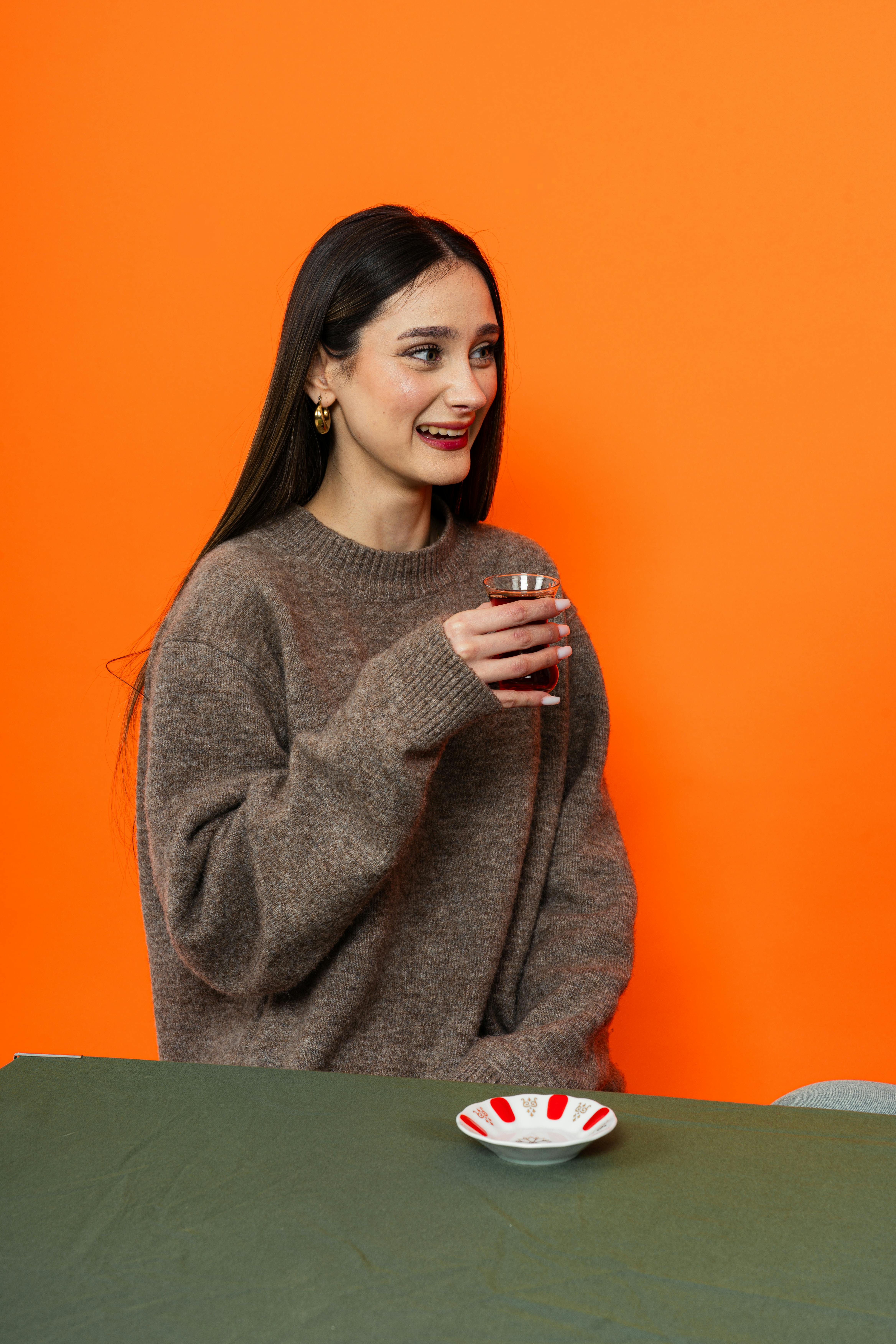 De franc Una dona jove amb un jersei còmode, somrient mentre sosté un got de te amb un fons taronja. Foto d'estoc