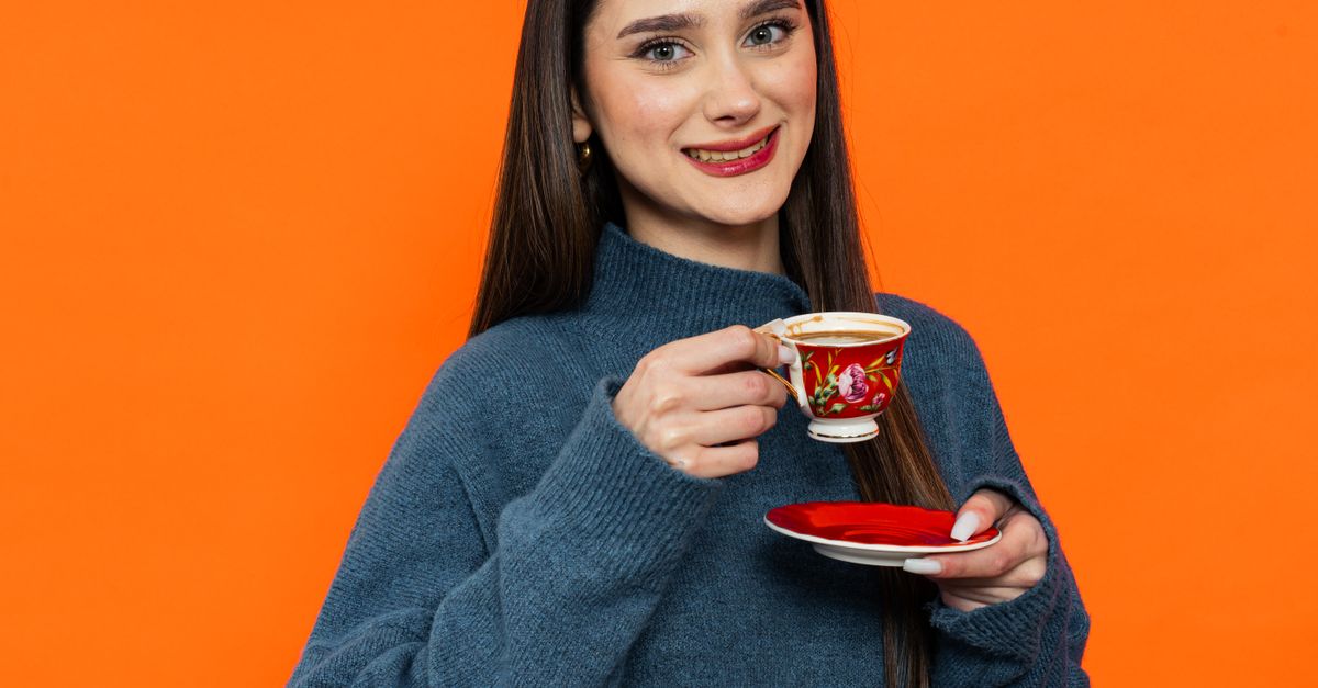 Young woman in Yalova enjoying tea against vibrant orange backdrop, wearing cozy sweater.