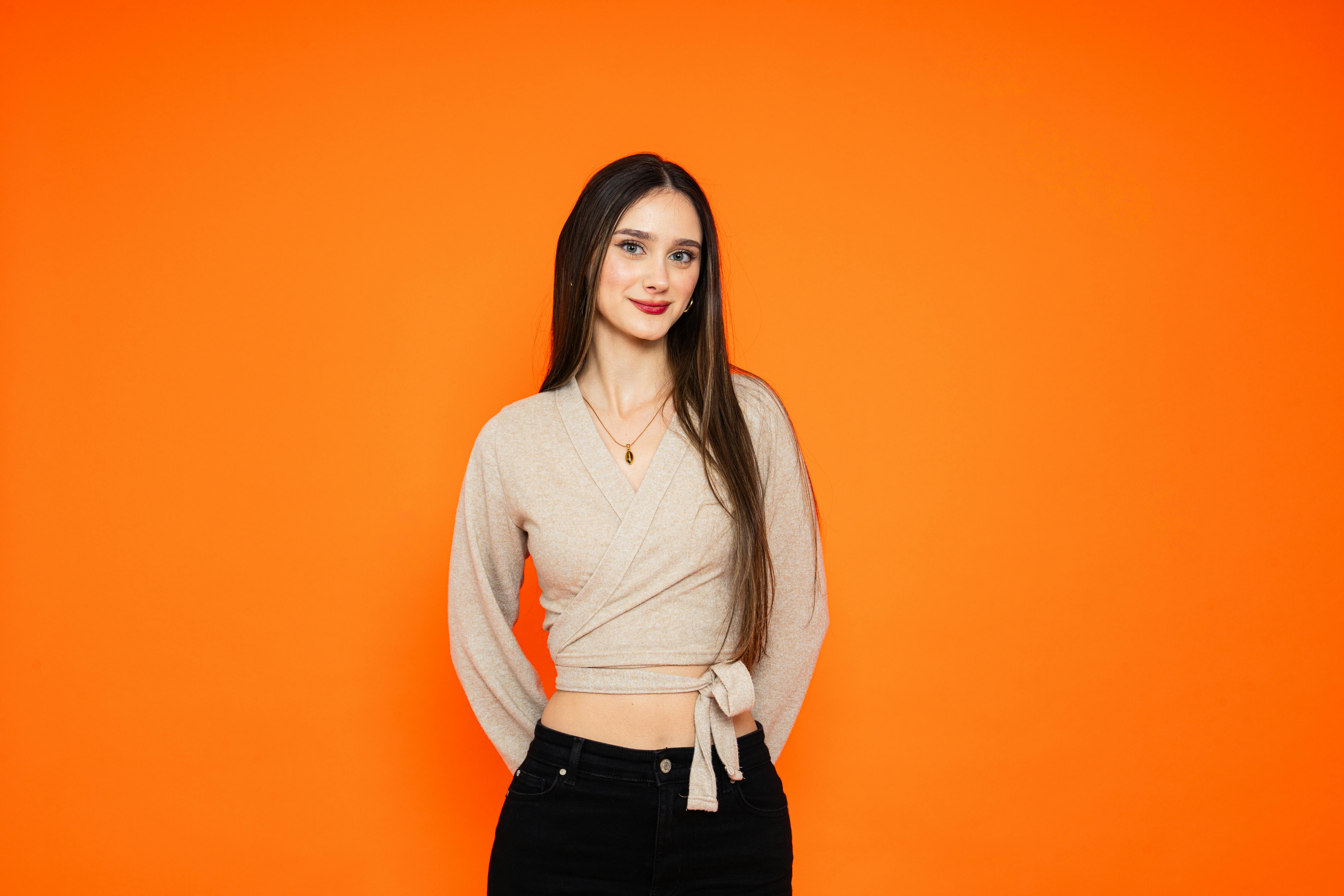 De franc Una dona jove amb els cabells llargs somrient amb confiança amb roba informal sobre un fons taronja vibrant. Foto d'estoc