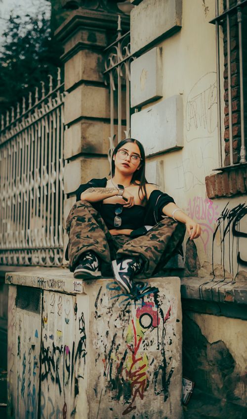 Free Stylish young woman in urban setting sitting on a graffiti wall wearing camo pants and glasses. Stock Photo