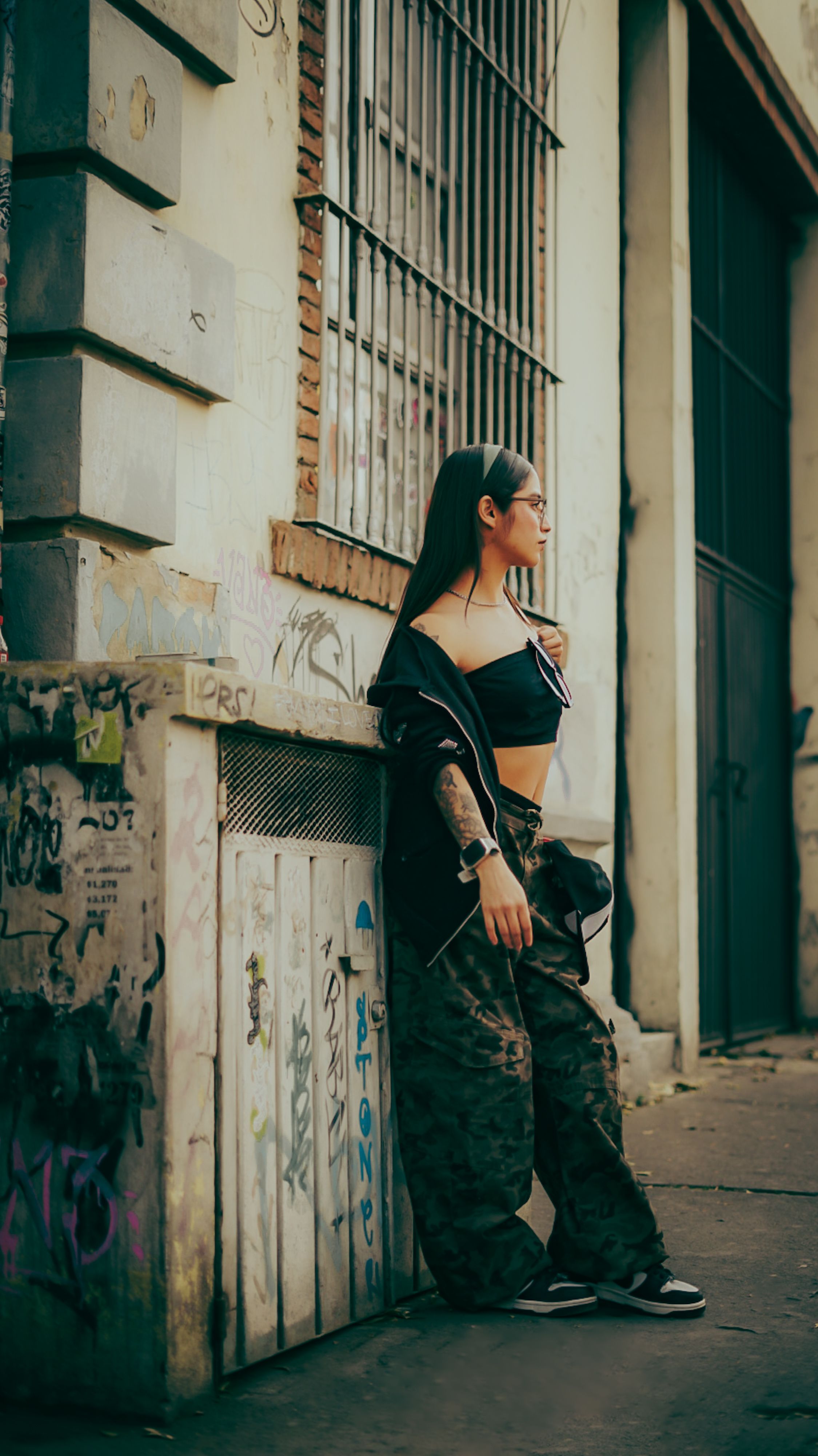 Jovem mulher com roupa moderna encostada em um muro grafitado em uma rua da cidade.