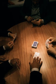 A gathering of adults playing cards around a wooden table in low, moody lighting.