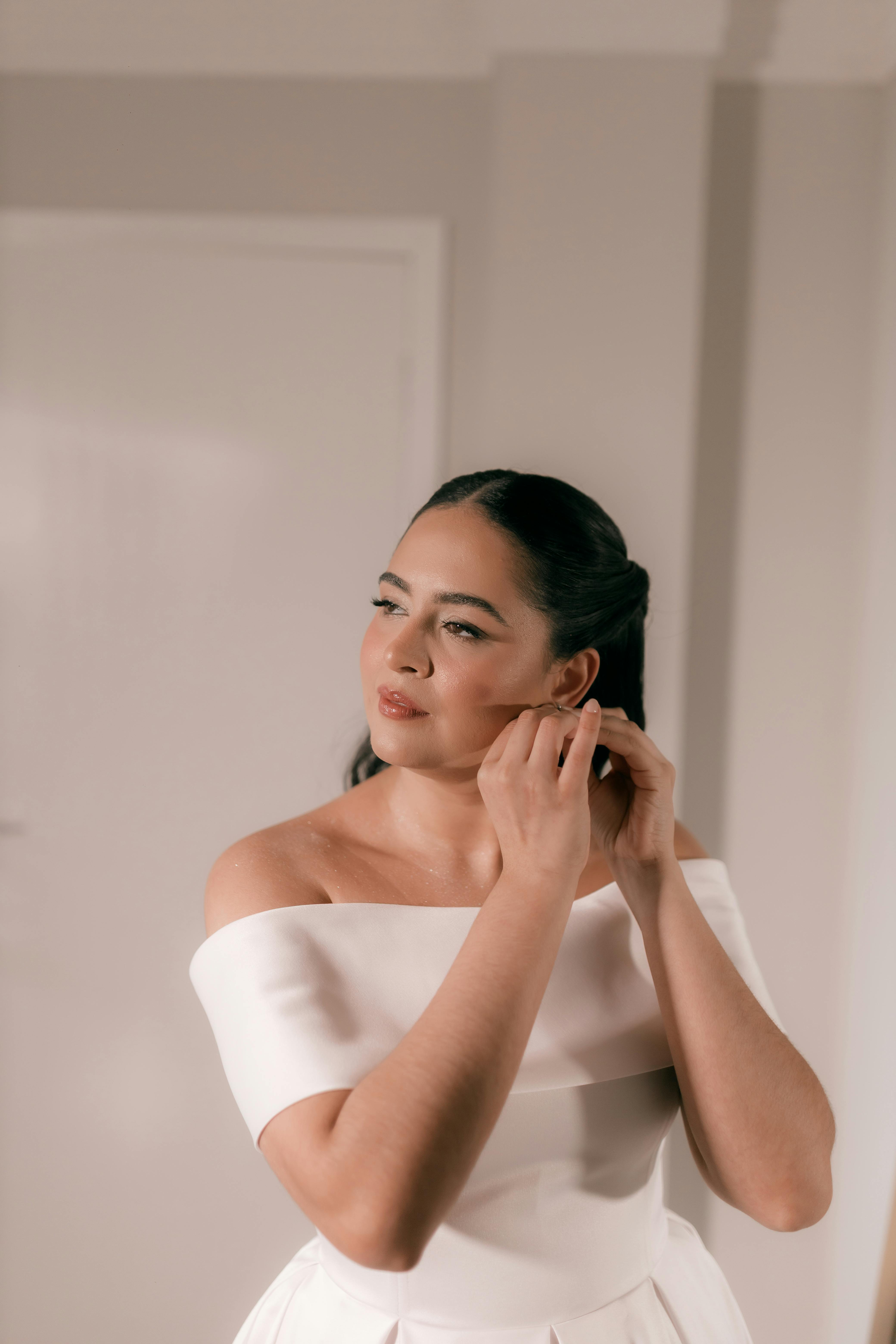 A bride elegantly adjusts her earring, dressed in a stylish white gown indoors.