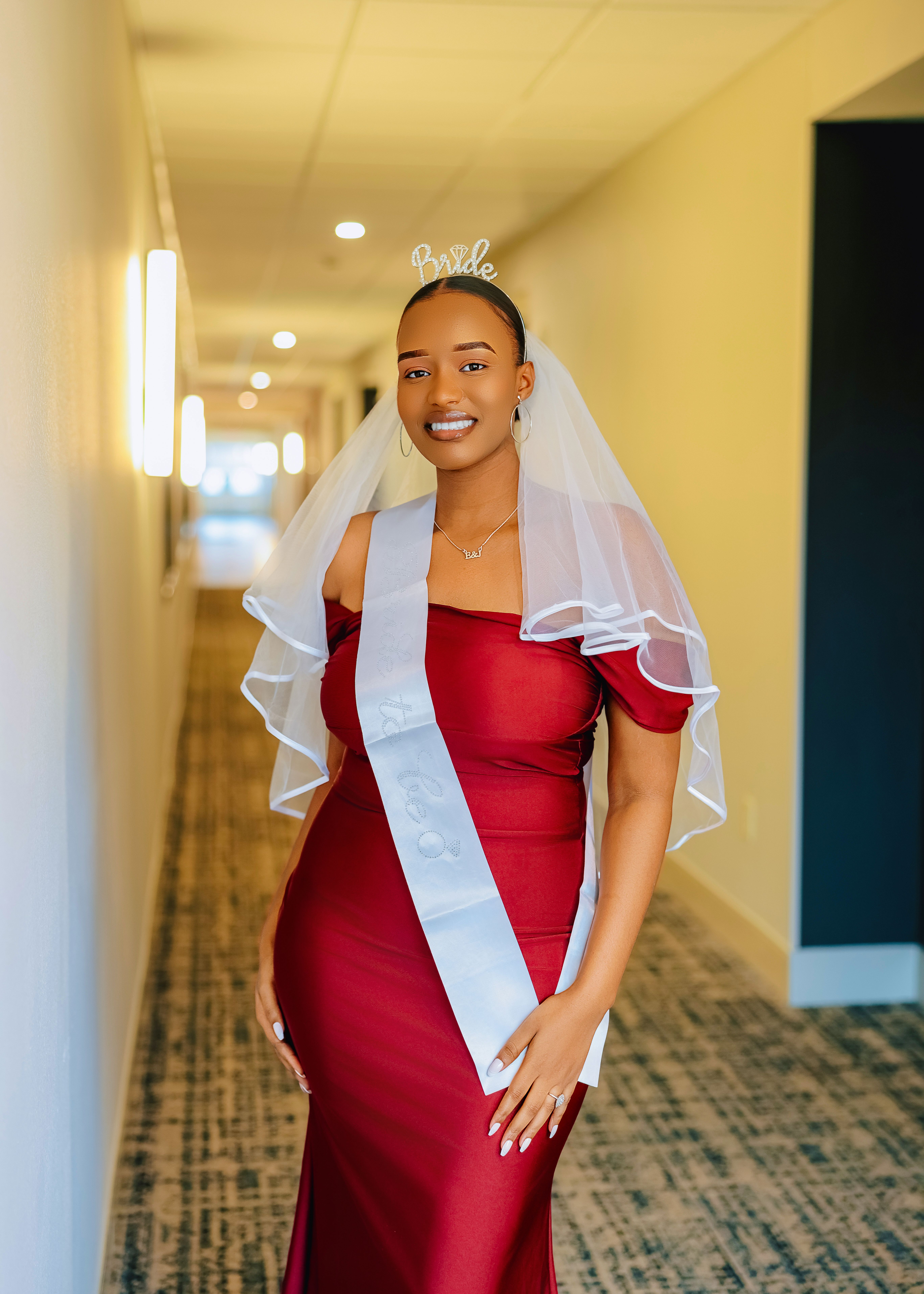 A smiling bride in a red gown with bridal sash and veil standing in a hallway.
