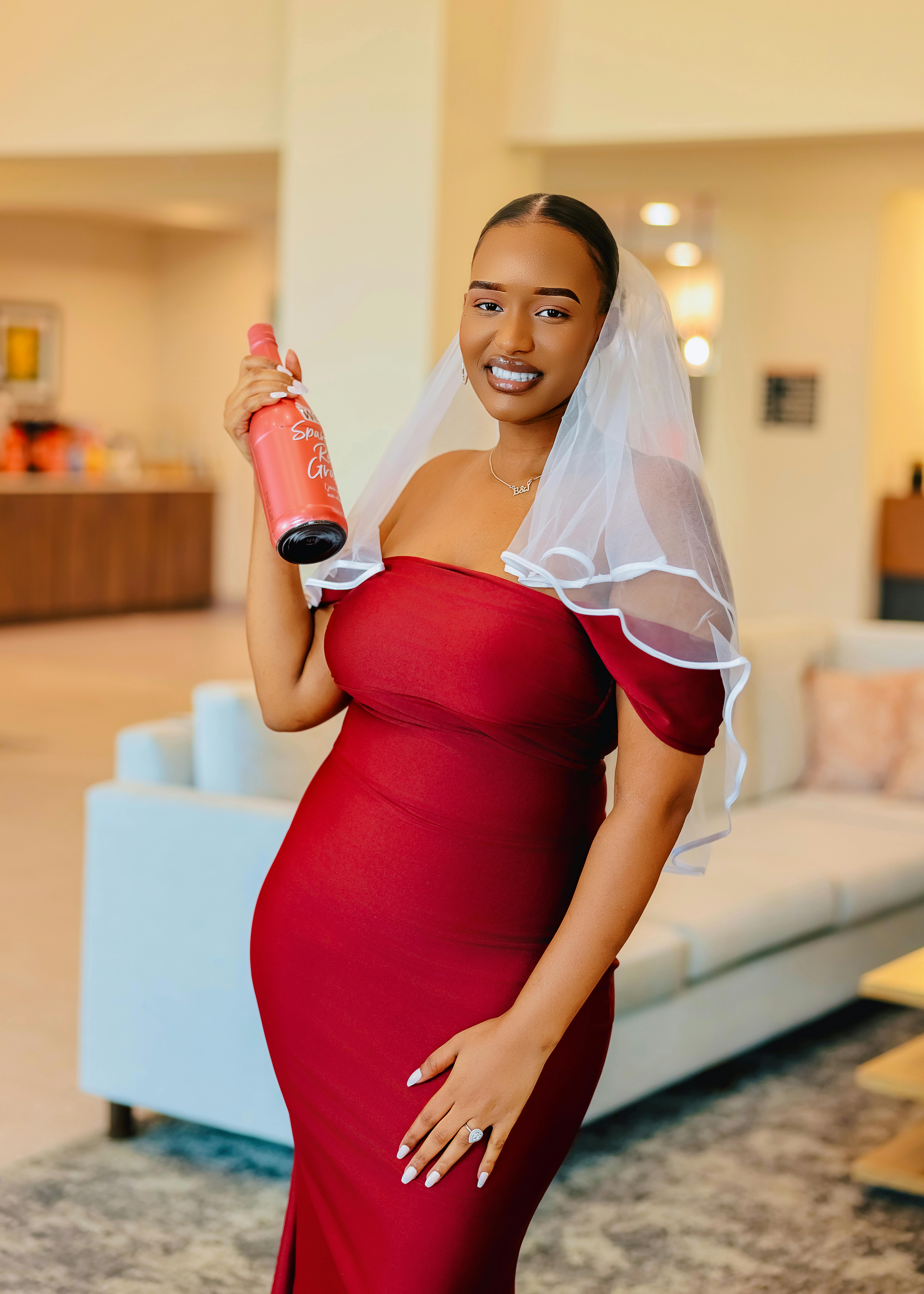 Joyful bride-to-be in red dress with veil holding a wine bottle indoors.