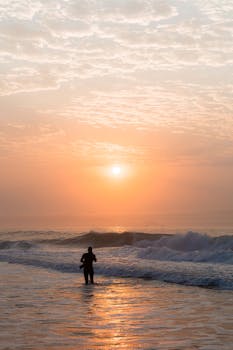 A lone silhouette stands by the ocean as the sun rises, casting a warm glow.