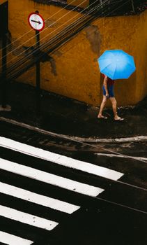 A person strolls on a wet city sidewalk under a blue umbrella beside a yellow wall.