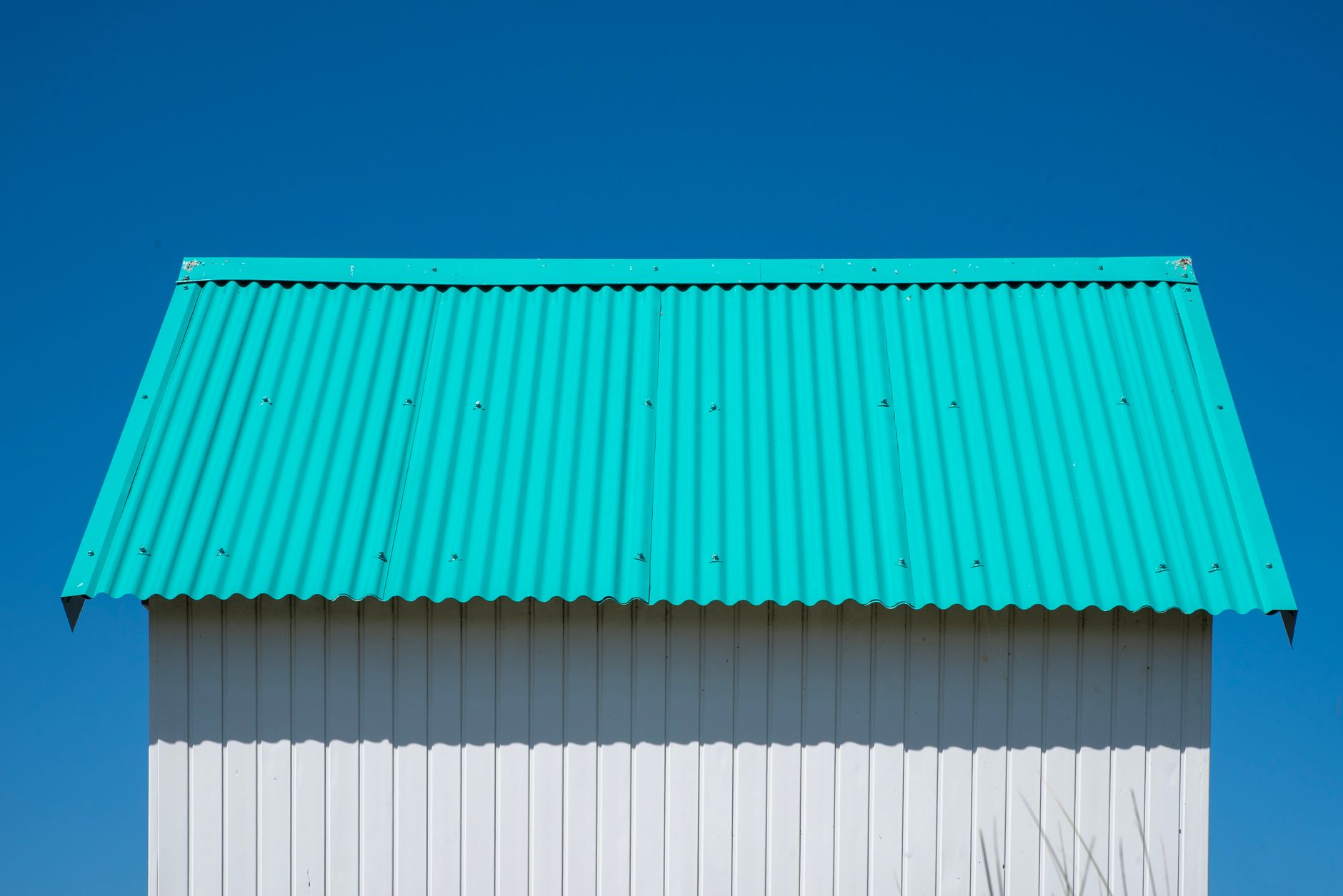 Simple architectural composition with a teal corrugated roof and white wall under clear blue sky.