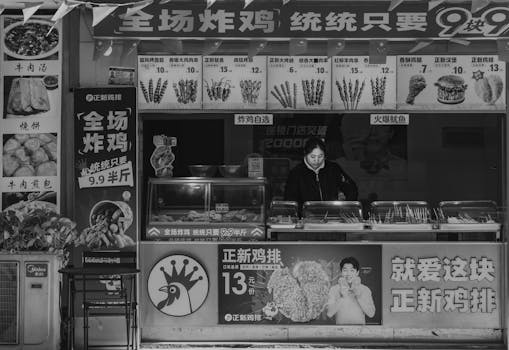 Black and white photo of a street food stall with vendor, displaying various menu items and prices.
