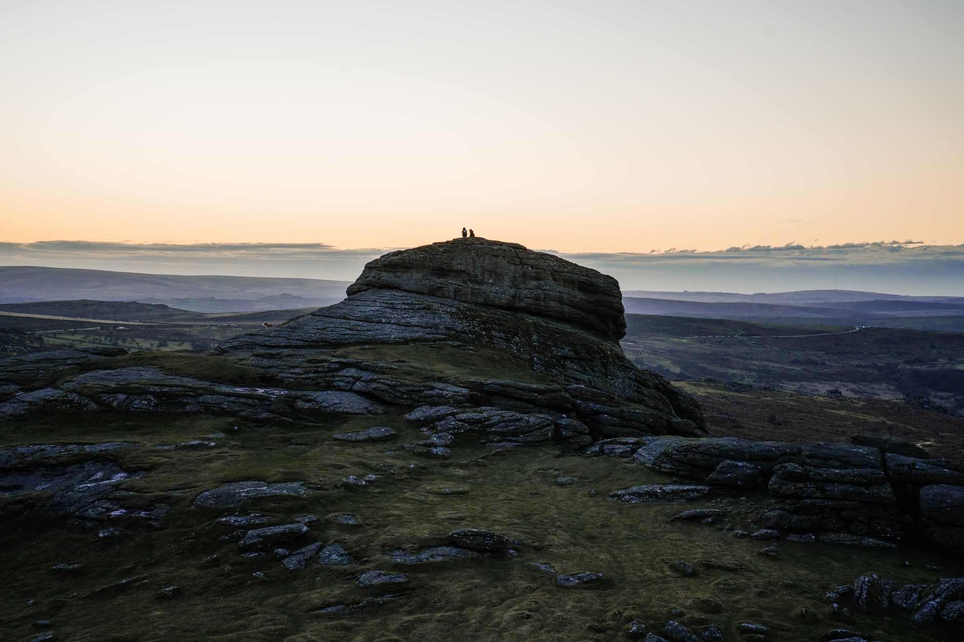 Two people on Haytor Rock with a scenic view at sunset in Dartmoor National Park, England.