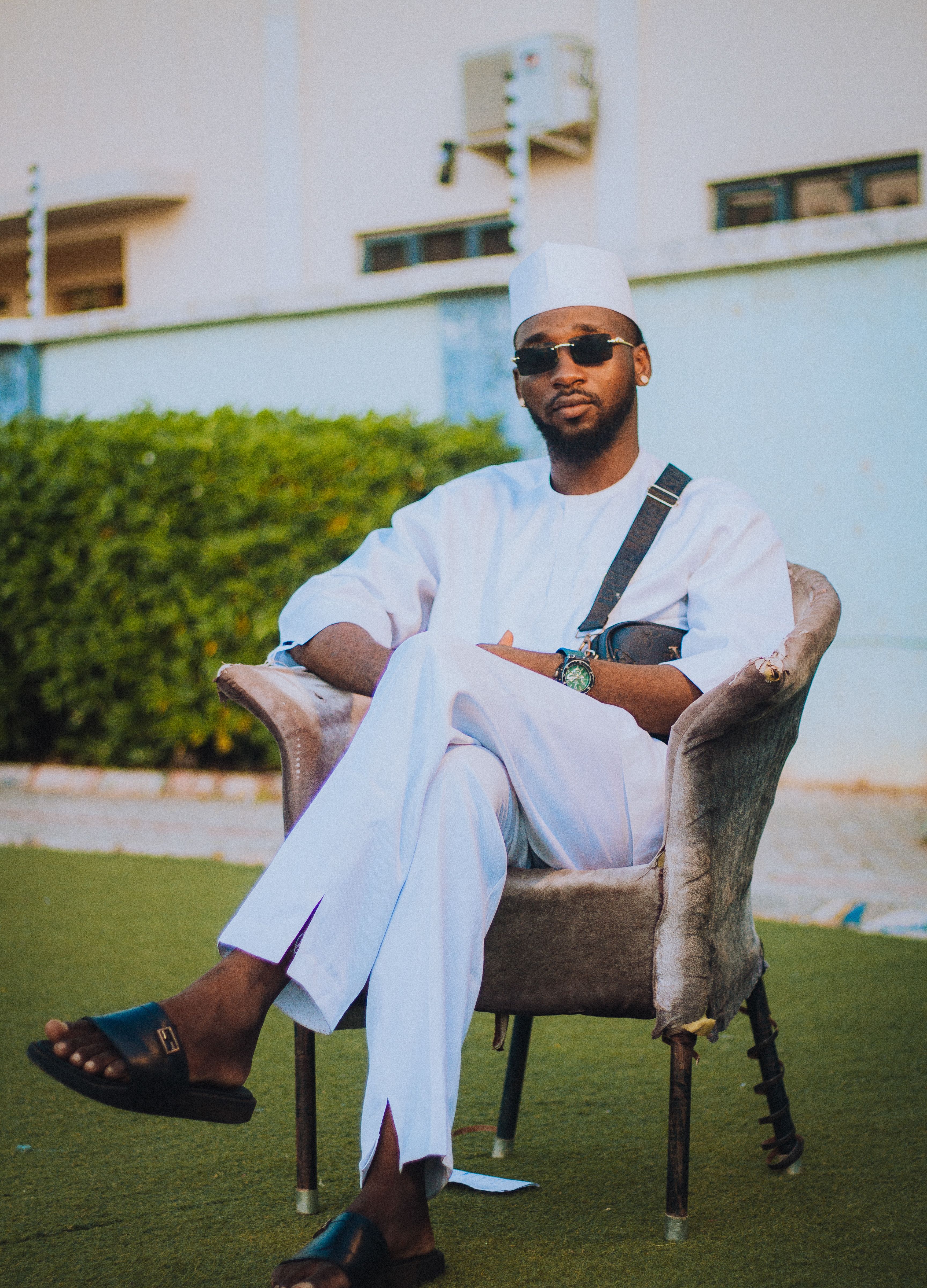 A fashionable man in traditional white clothing and sunglasses seated outdoors on a sunny day.