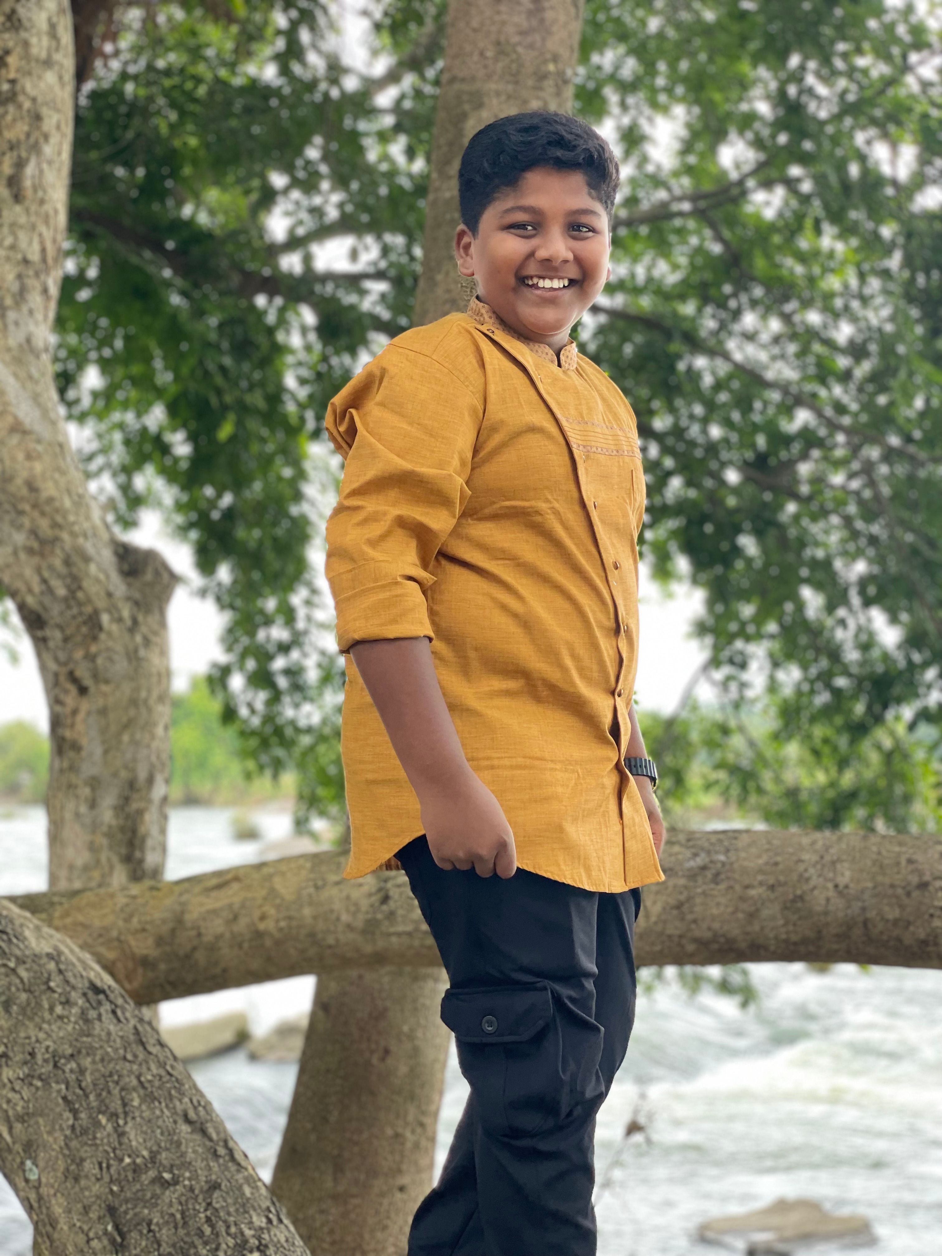 A cheerful boy in a yellow shirt poses on a tree in Srirangapatna, India.