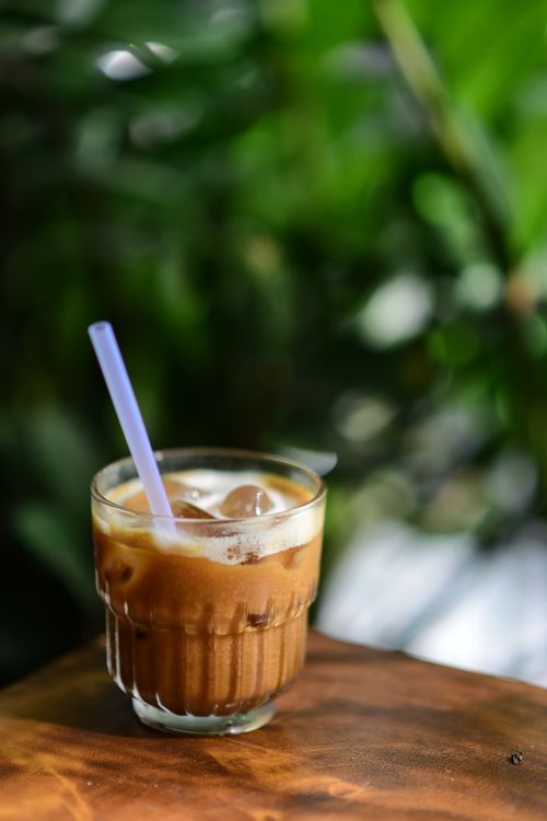 Free A glass of iced coffee with a straw sits on a wooden surface, surrounded by vibrant green leaves. Stock Photo