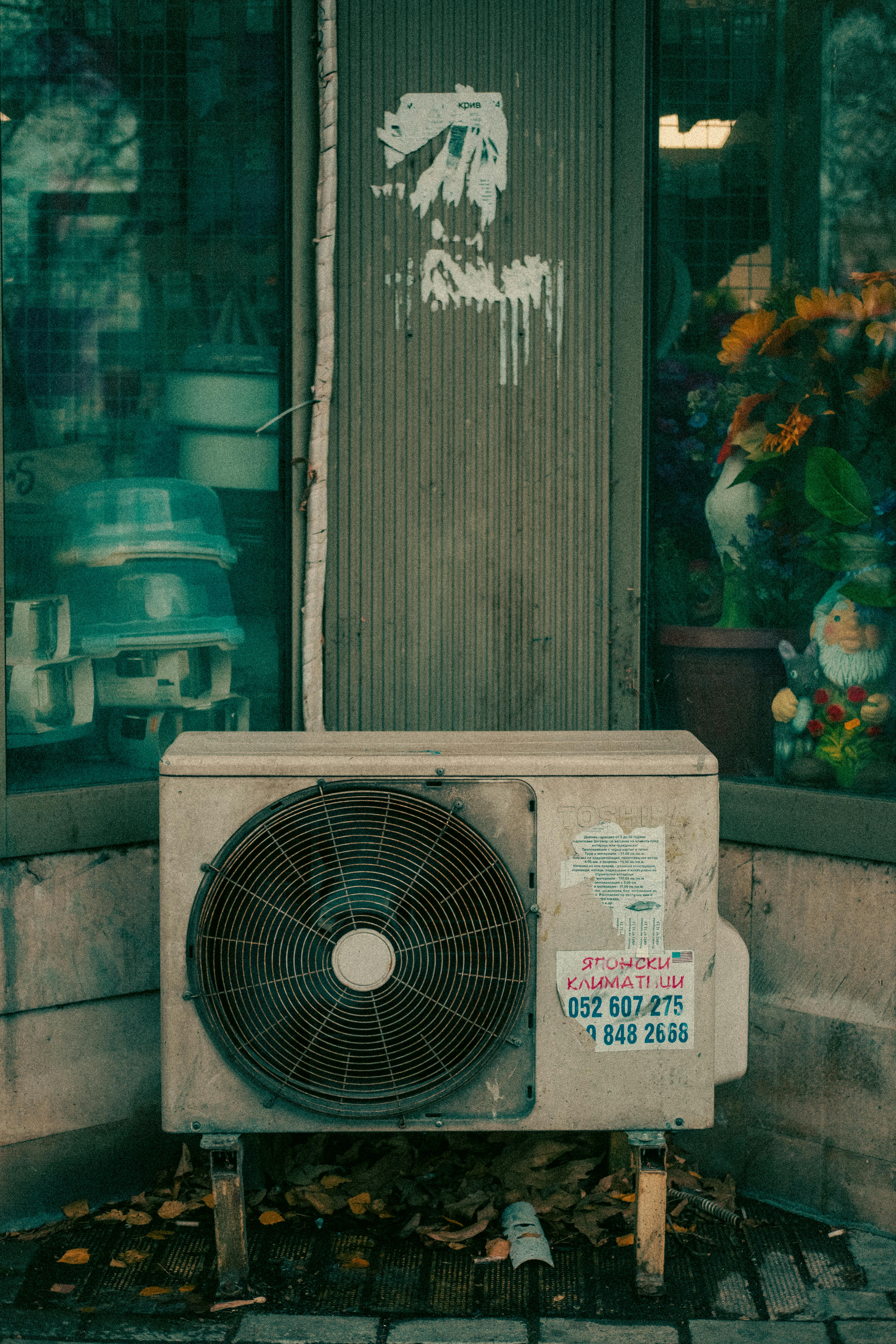 Aged air conditioning unit outside a Varna shop displaying flowers and garden decor.