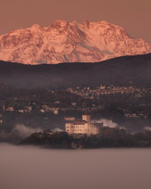Free A breathtaking view of a castle nestled below snow-capped mountains at sunrise, surrounded by a misty landscape. Stock Photo