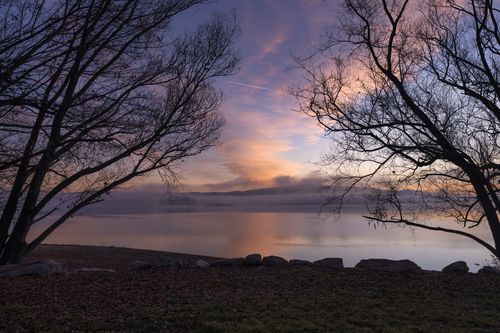 Free A breathtaking sunset over a calm lake, framed by silhouetted trees, creating a tranquil landscape scene. Stock Photo