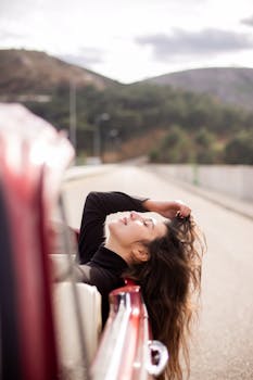 Woman relaxing out of a red car, enjoying the breeze on a scenic mountain highway.