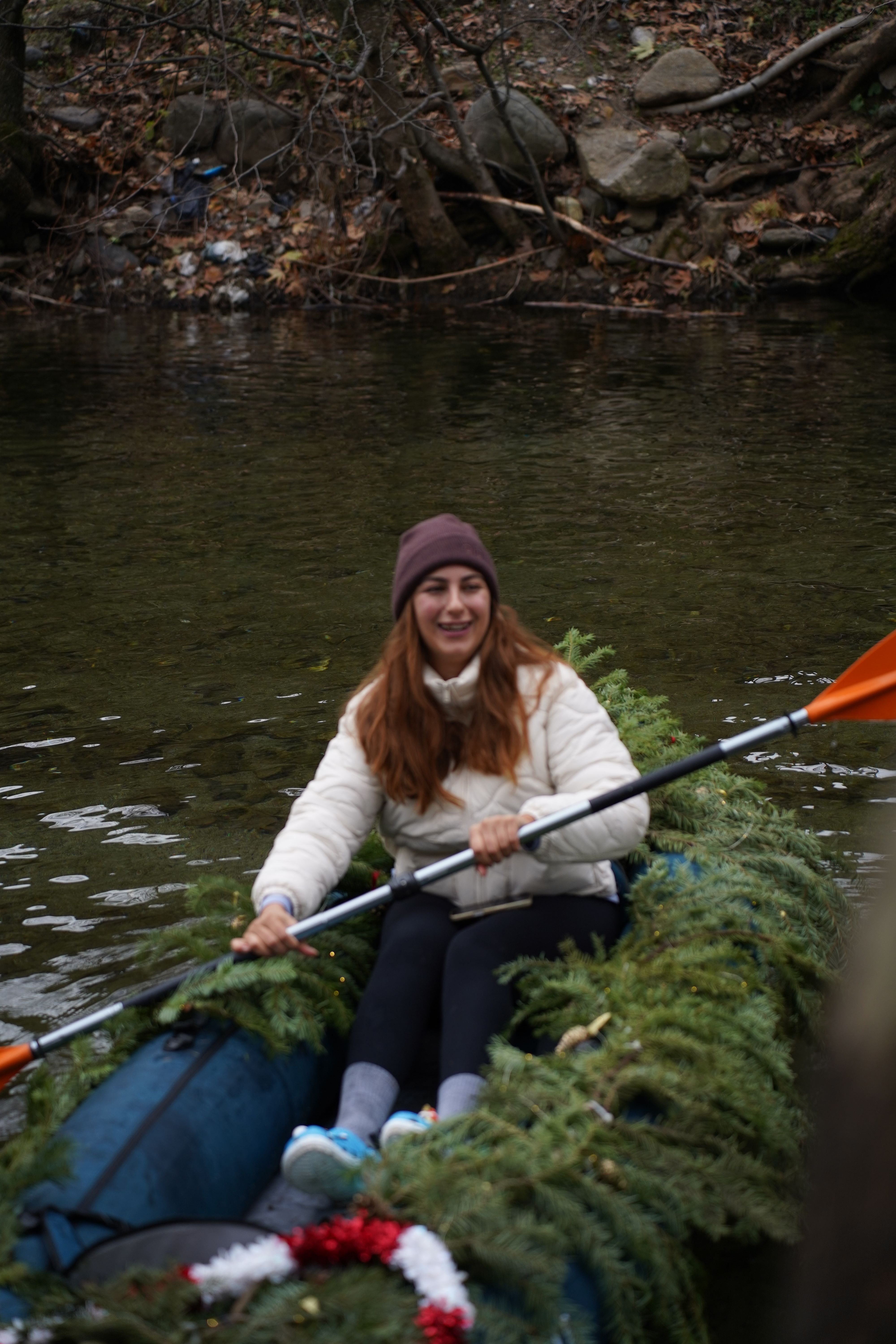 A woman paddles a canoe decorated with greenery on a serene river in a winter forest setting.