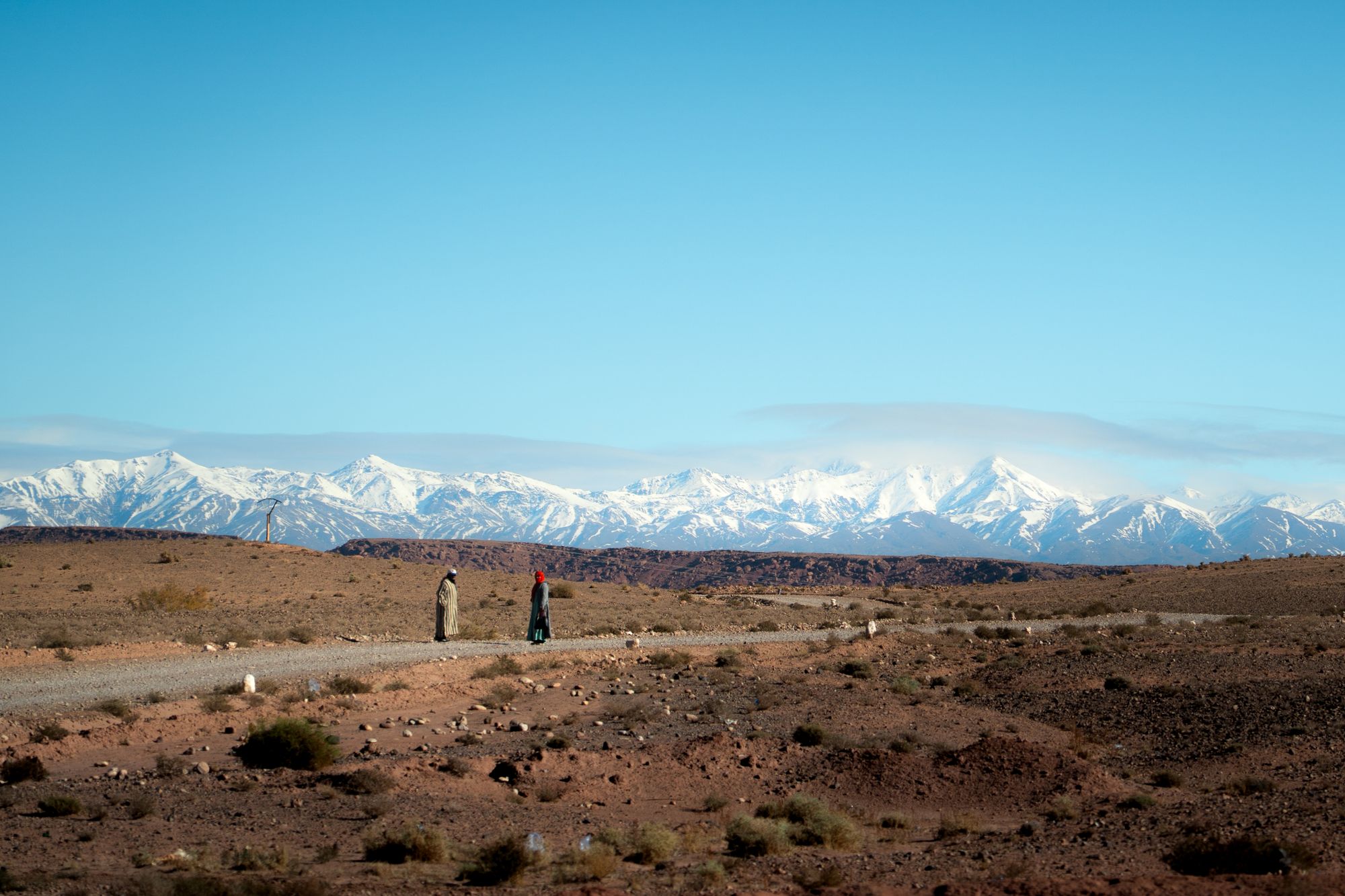 Travellers crossing a broad landscape beneath the snow-capped Atlas Mountains