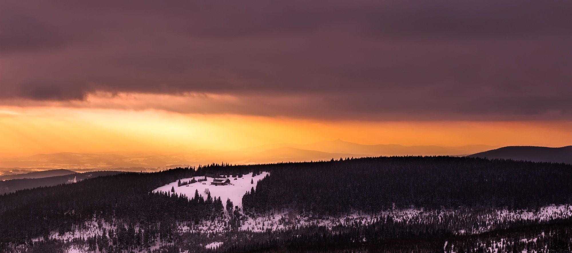 Breathtaking winter sunset over snow-covered hills and dense forest with a dramatic sky.
