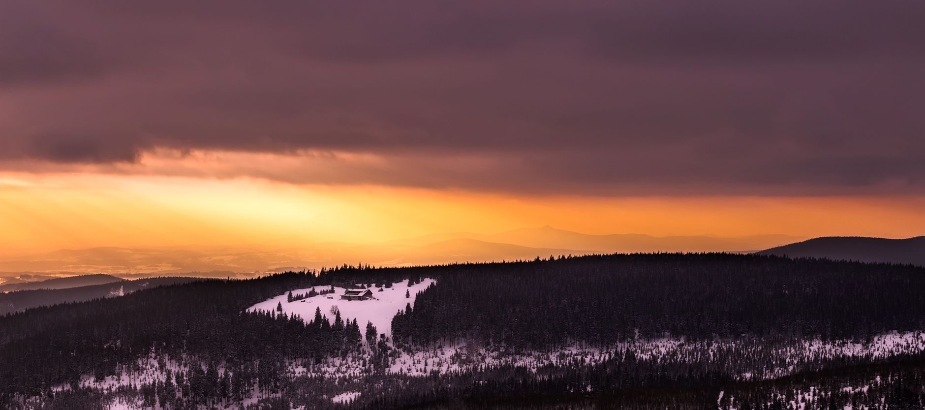 Breathtaking winter sunset over snow-covered hills and dense forest with a dramatic sky.