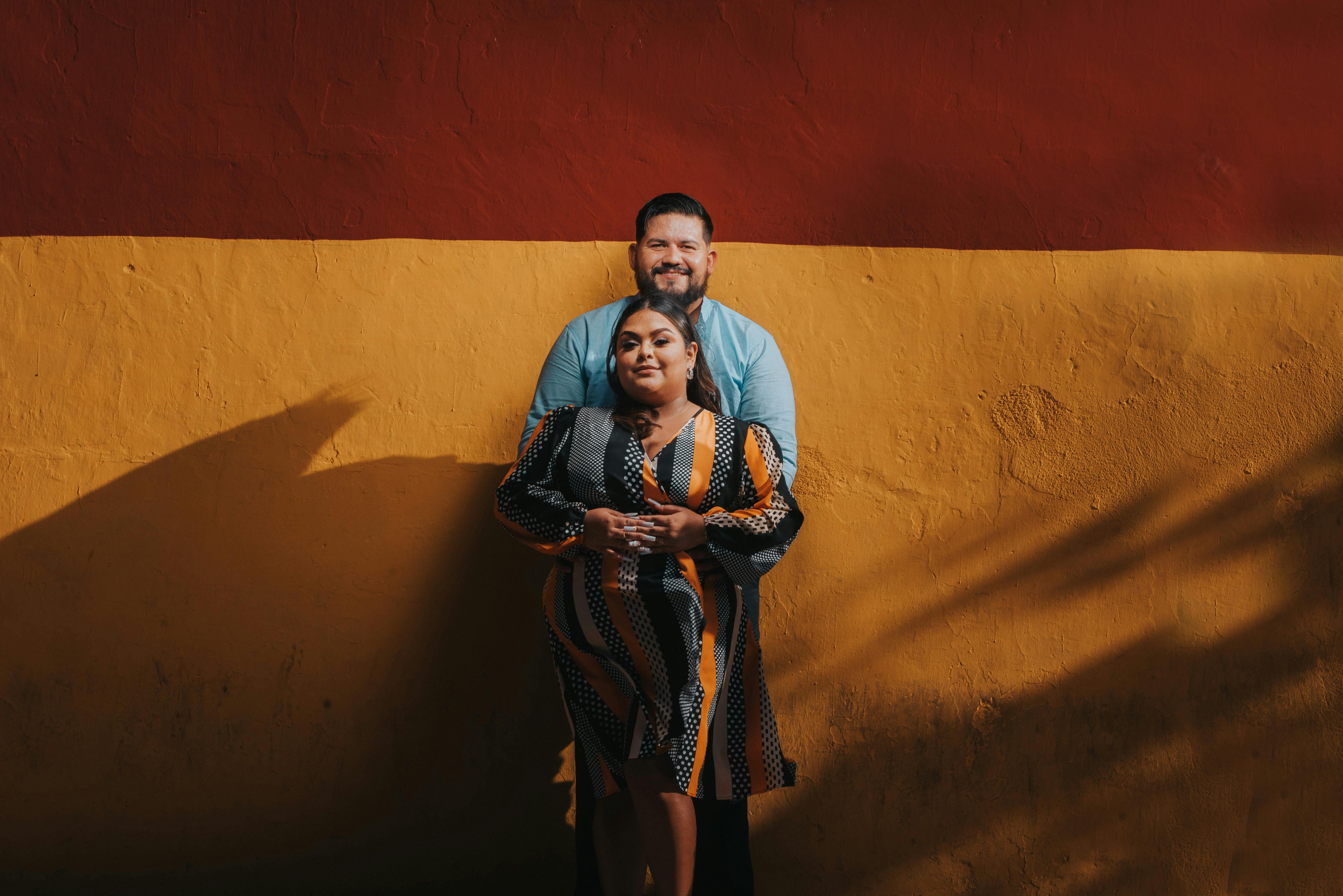 Gratuit Un couple se tient côte à côte devant un mur rouge et jaune éclatant, enlacé. Photos