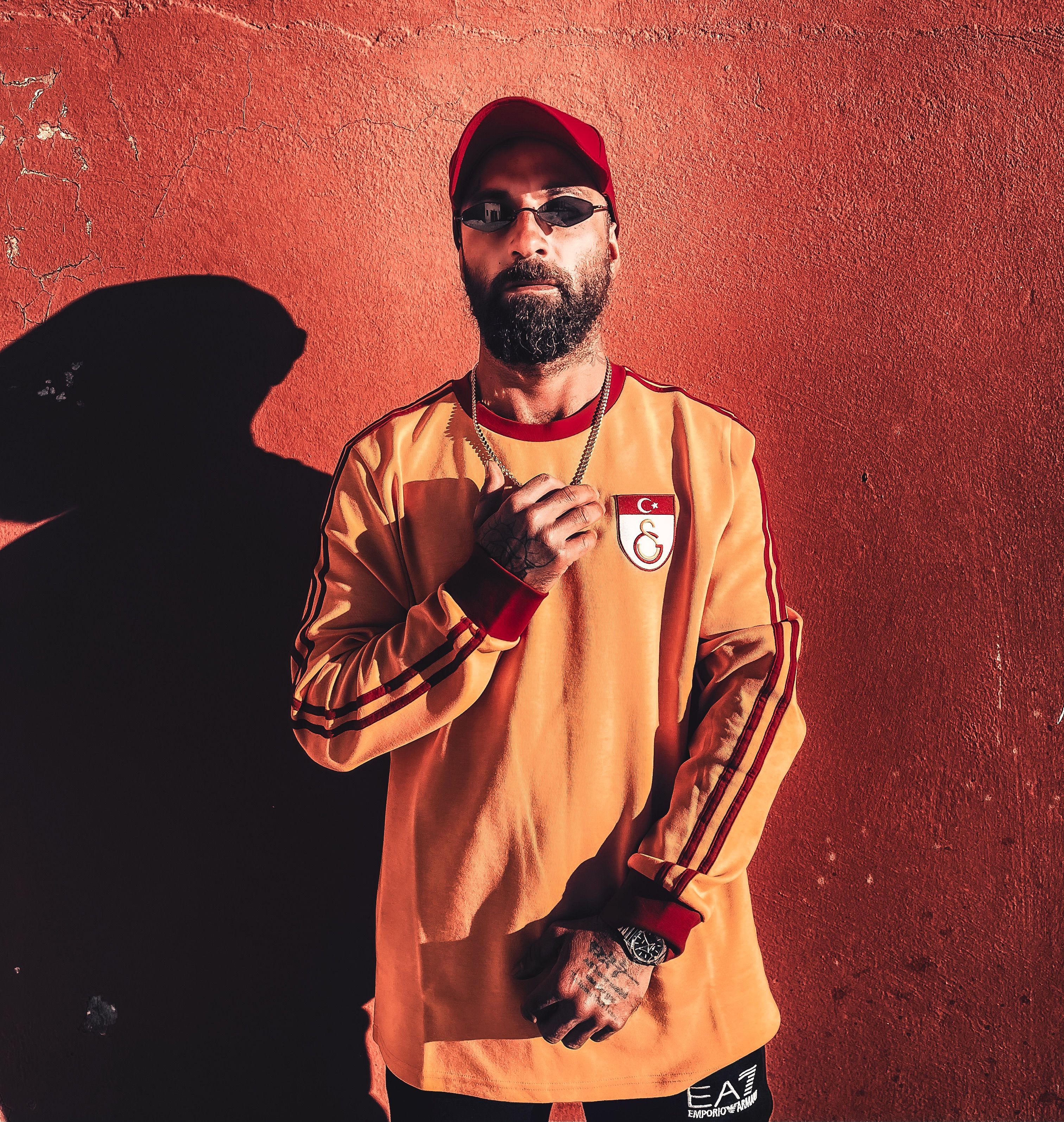 Confident man in sunglasses and bright jersey poses against a textured wall, showcasing bold street fashion.