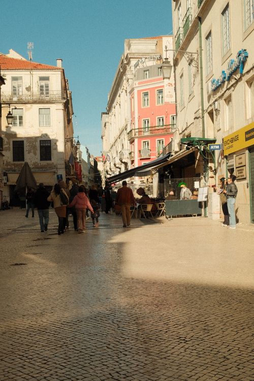 Free Charming Lisbon street with colorful buildings, cafes, and people enjoying a sunny day in Portugal. Stock Photo