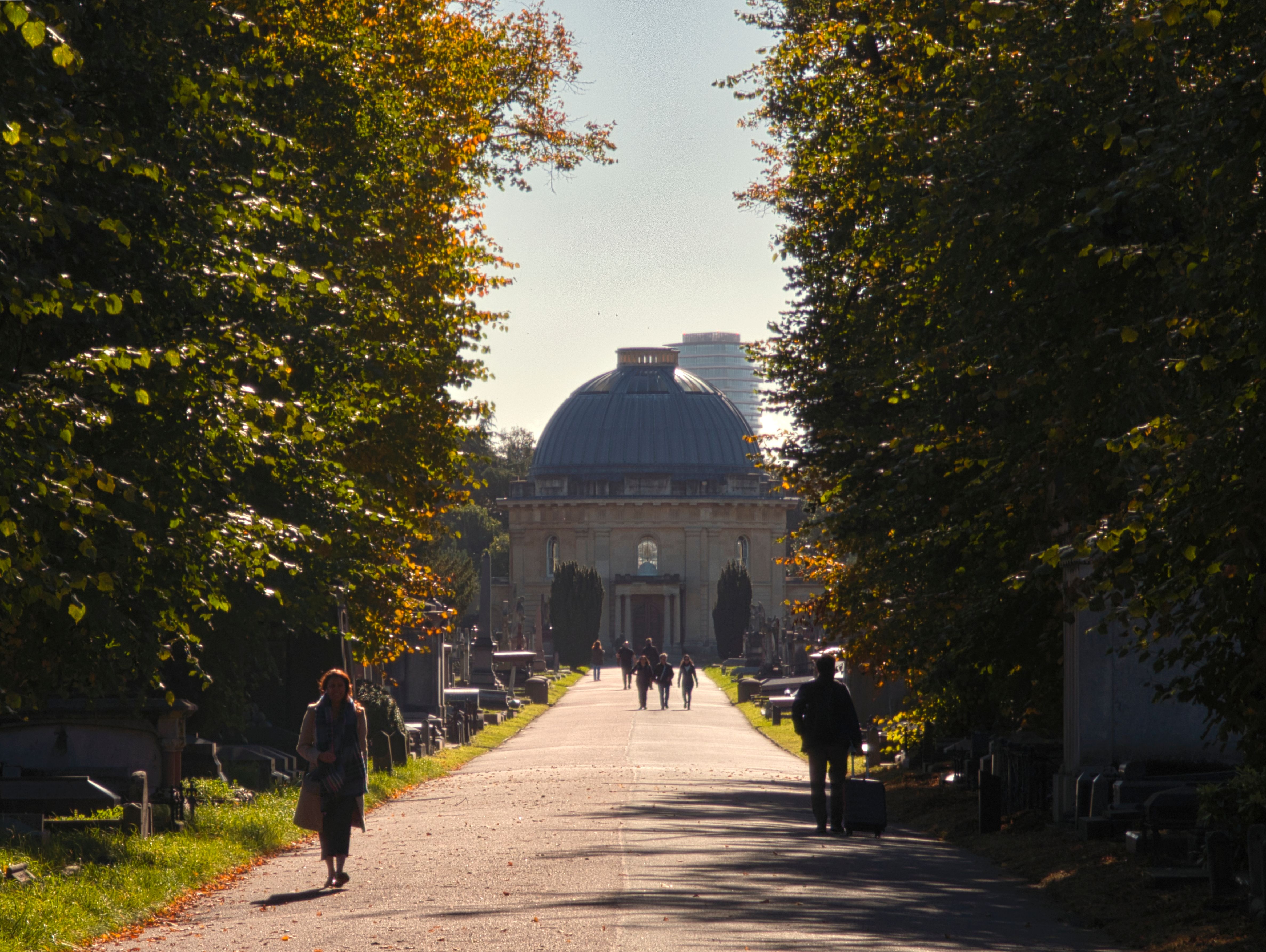 Visitors walking through a sunlit pathway at Brompton Cemetery, London, surrounded by autumn foliage.