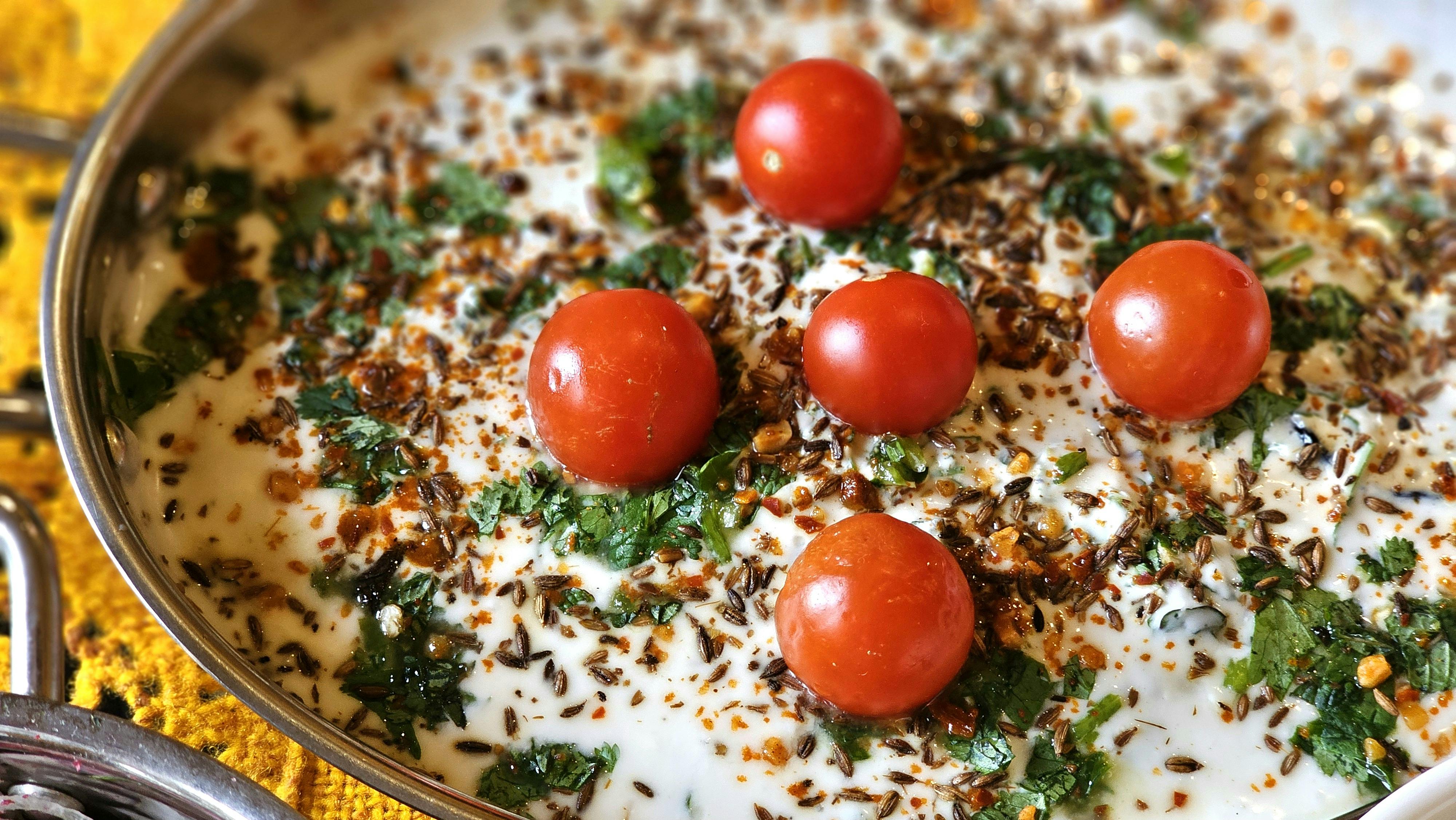 Close-up of creamy raita garnished with cherry tomatoes, spices, and fresh herbs in a stainless steel bowl.
