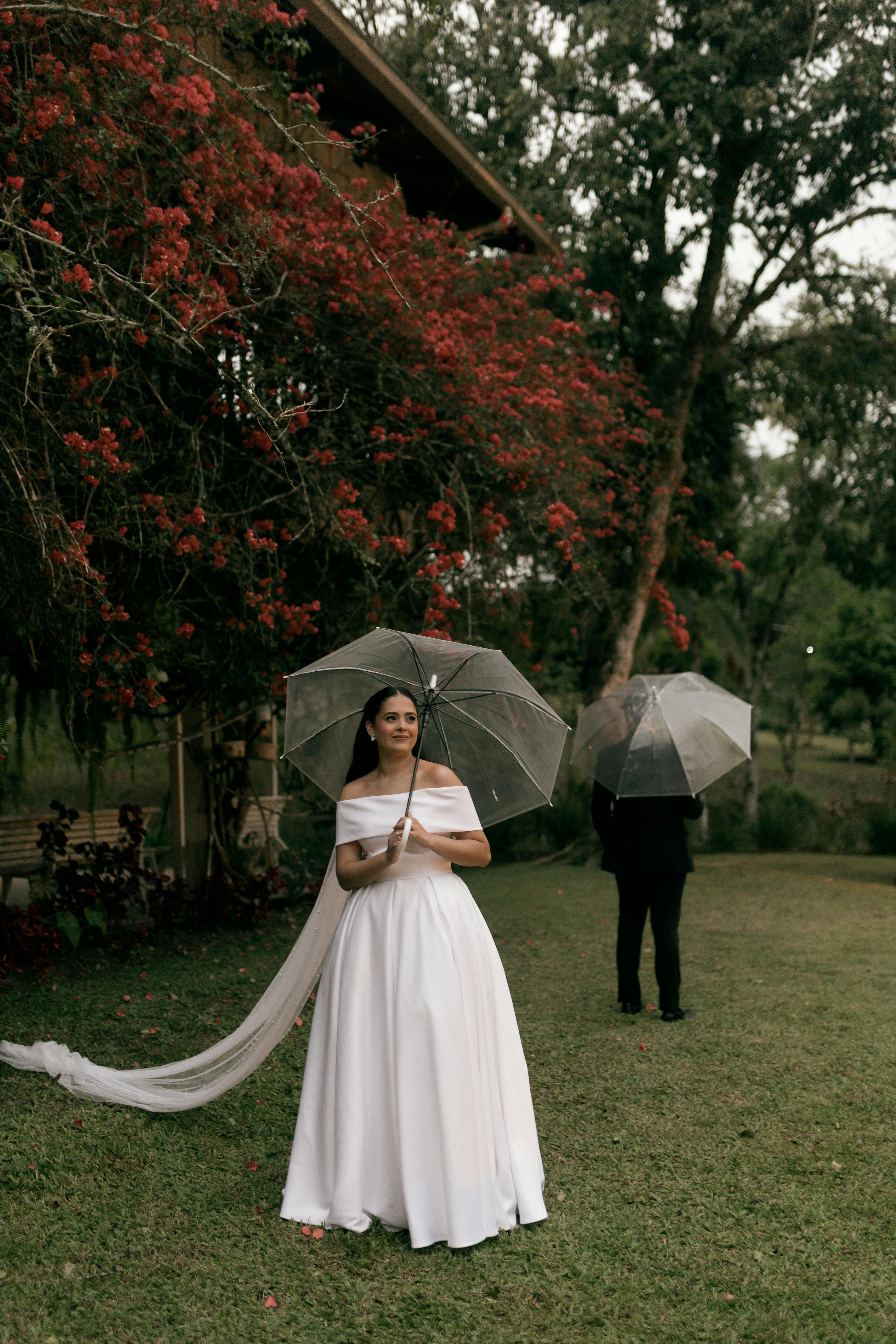 Gratuit Une mariée tenant un parapluie à l'extérieur par temps nuageux, entourée de verdure et de fleurs rouges. Photos