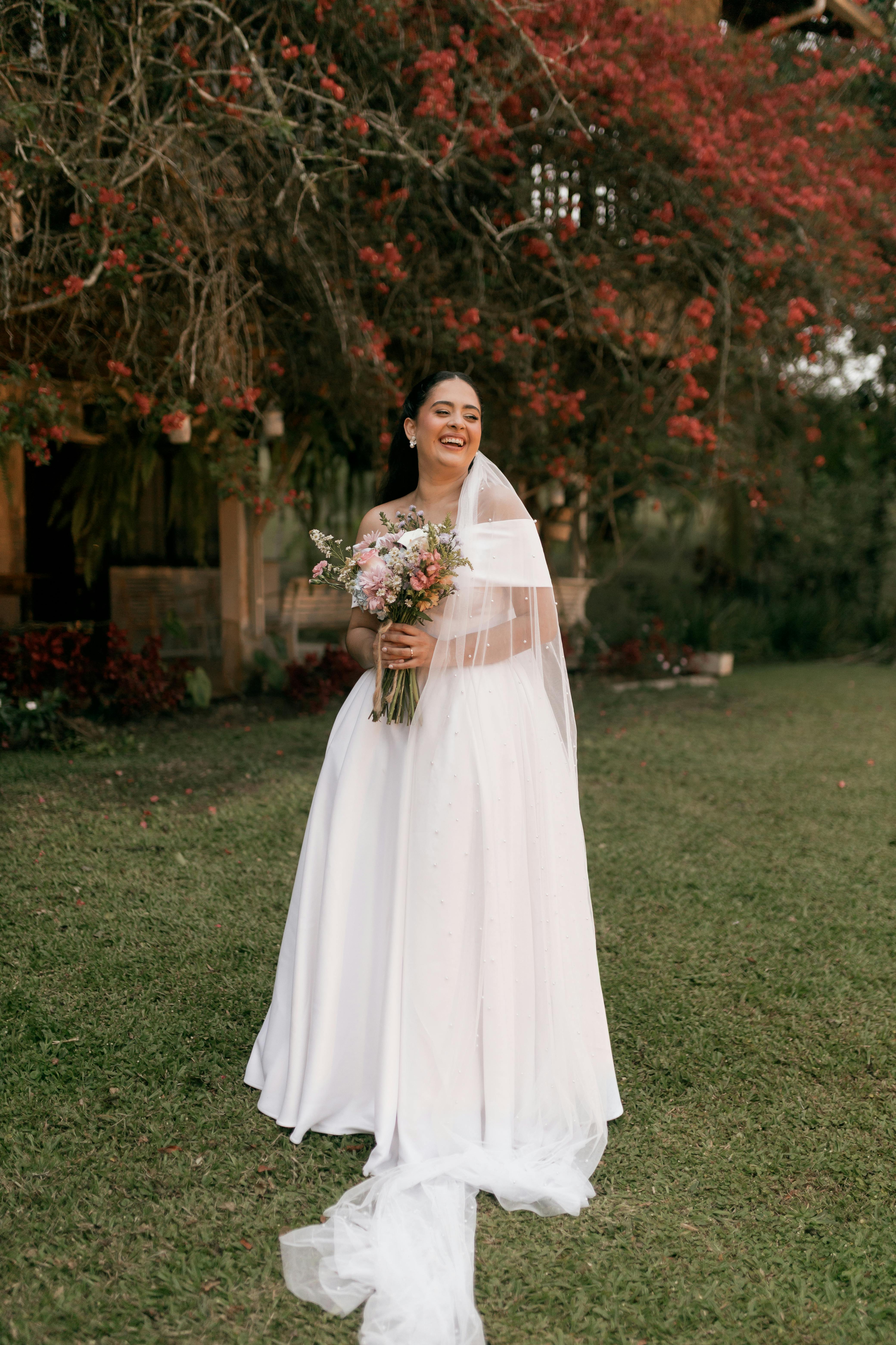 Gratuit Une mariée souriante en robe blanche se tient à l'extérieur, tenant un bouquet de fleurs. Photos