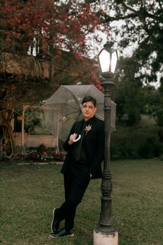 Man in black suit with transparent umbrella leaning on a lamp post in a garden setting.
