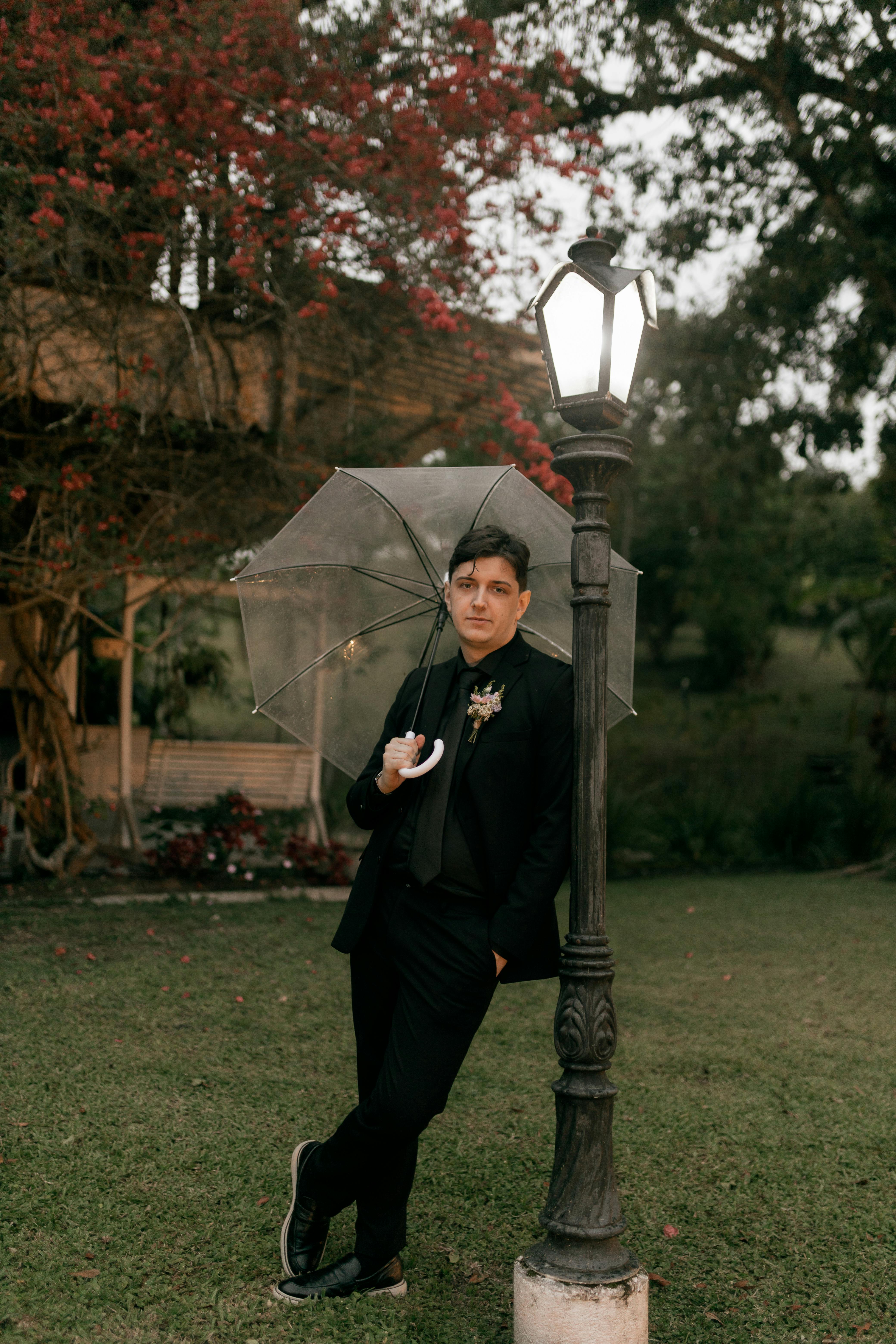 Gratuit Un homme en costume noir, parapluie transparent à la main, s'appuie contre un lampadaire dans un jardin. Photos