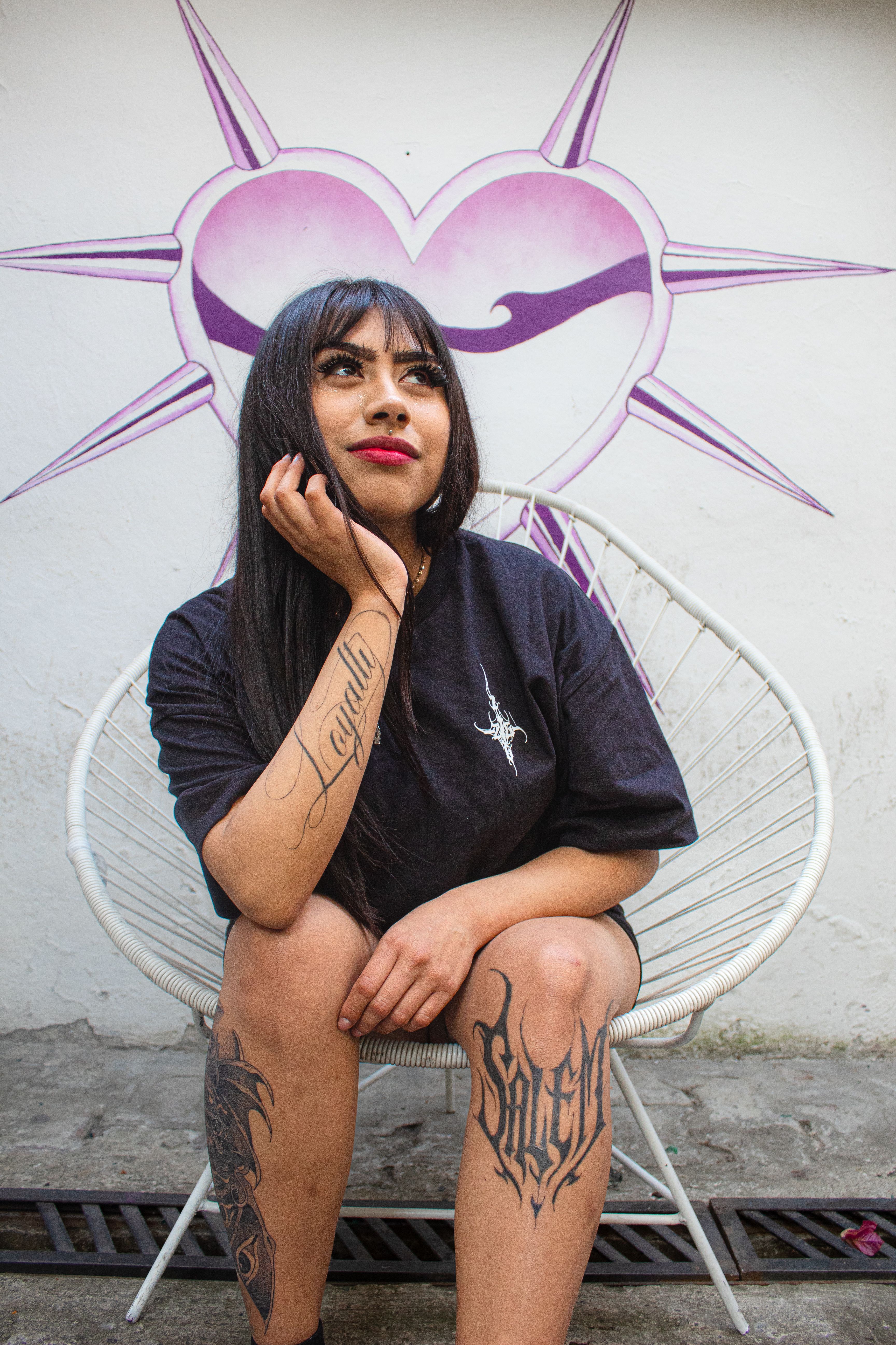 Young woman with tattoos sitting in a chair against a heart mural background.