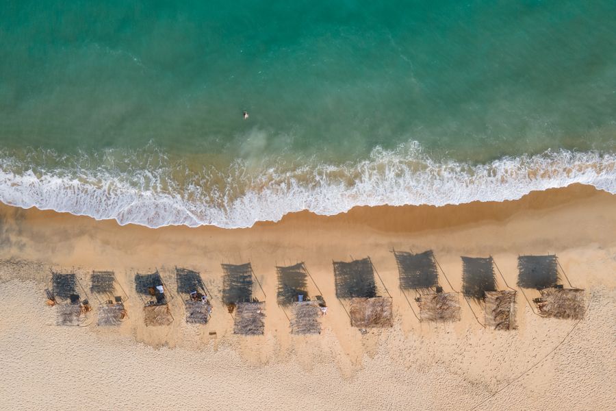 Aerial view of straw huts casting shadows on the sandy beach of Trancoso, Bahia, Brazil.