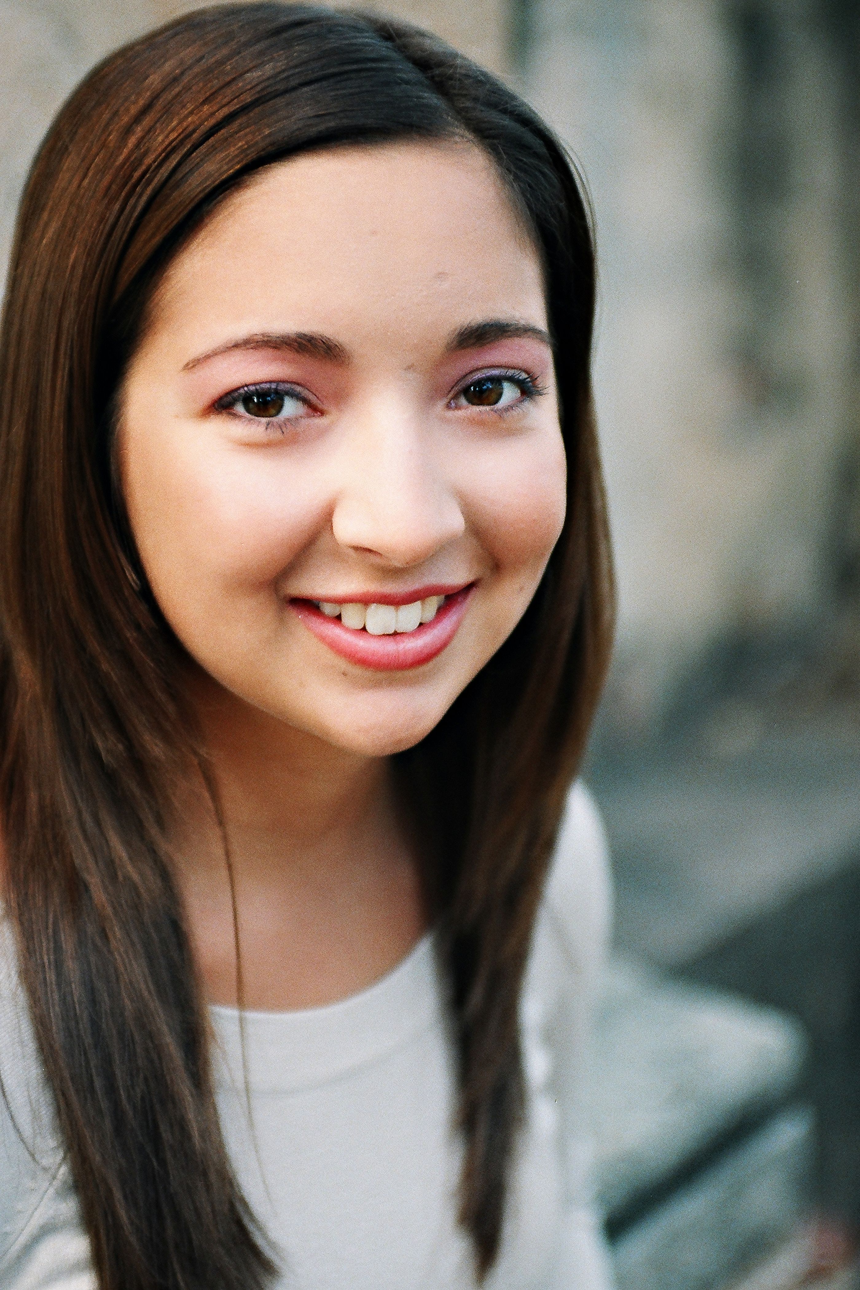 A young woman with straight hair smiles warmly in an outdoor setting, exuding natural beauty and charm.