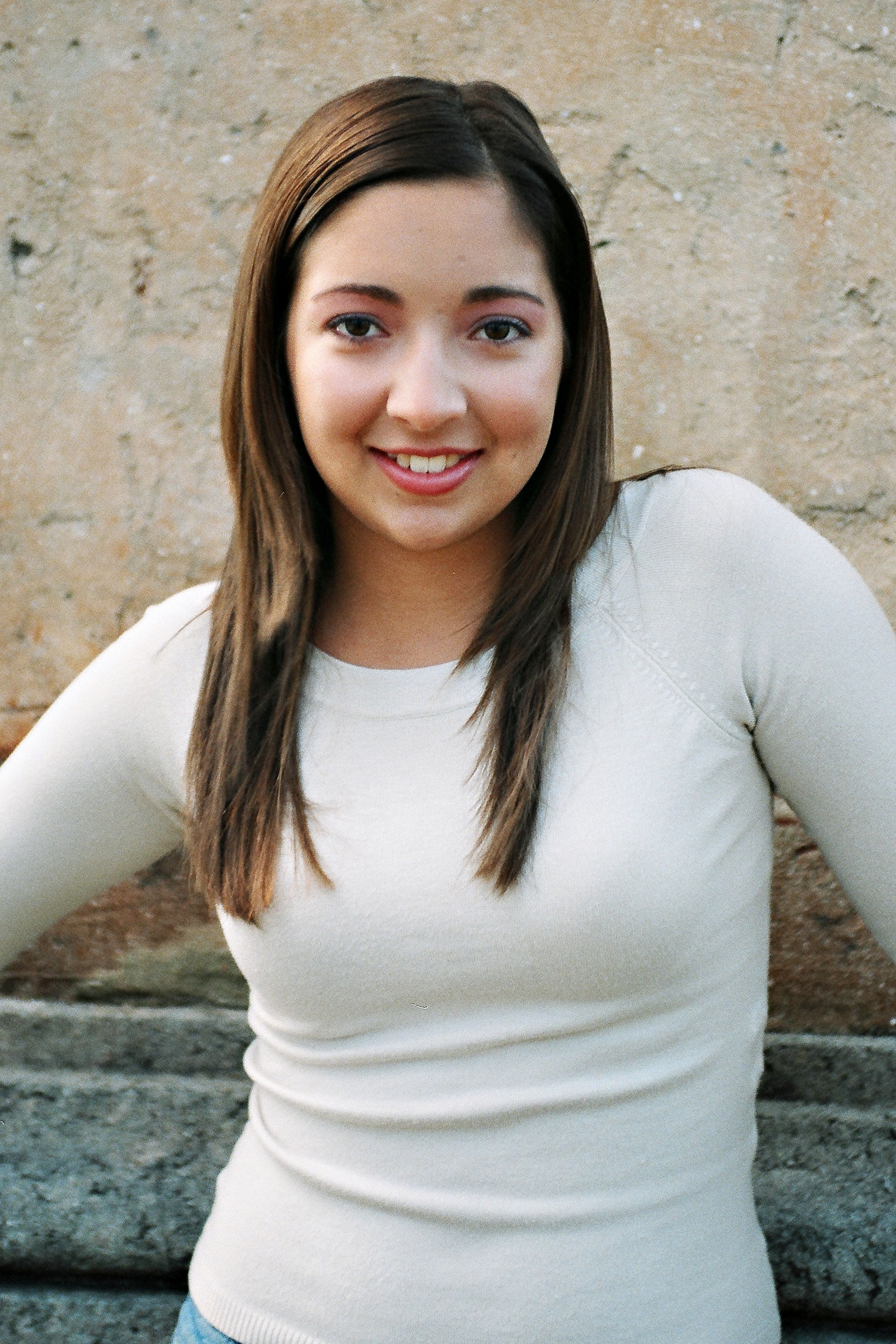 Smiling young woman in casual attire posing against a rustic stone wall outdoors.