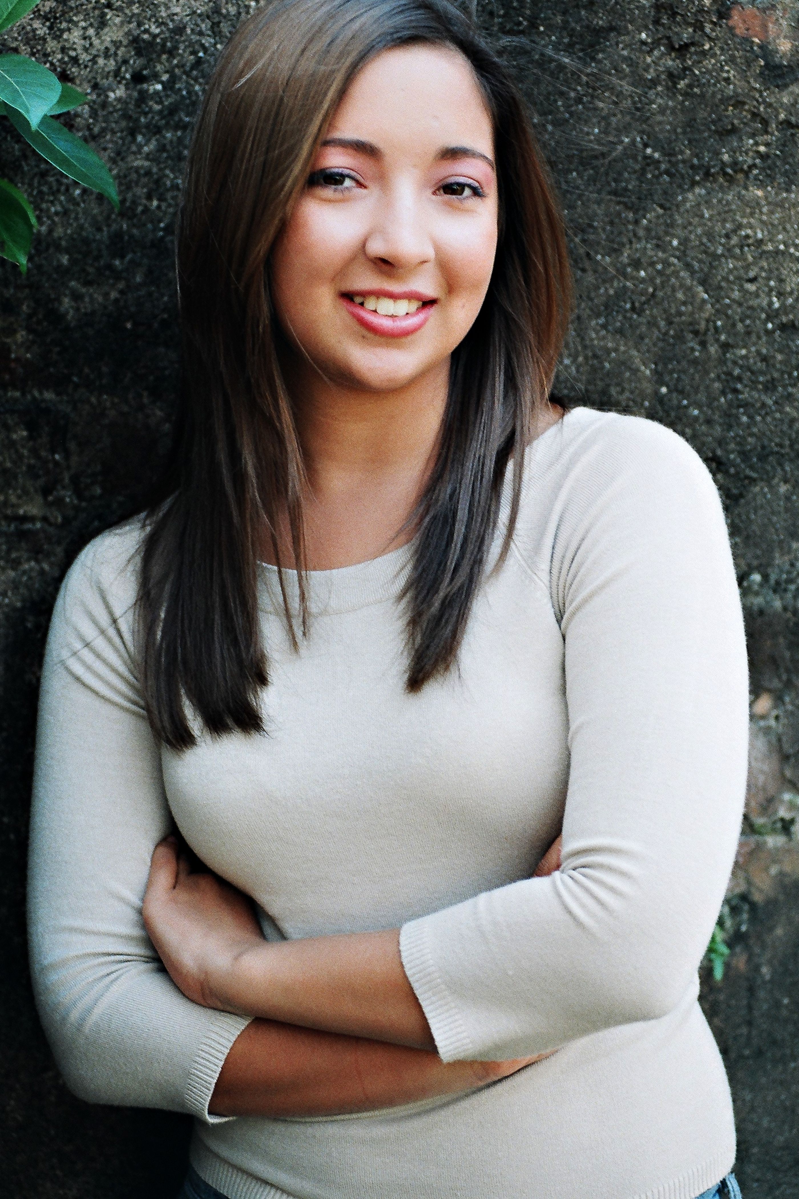A friendly young woman poses confidently against a rustic wall, exuding warmth and charm.