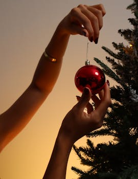 Close-up of hands placing a red ornament on a Christmas tree, creating a warm festive atmosphere.