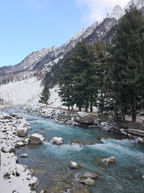 Free Serene winter scene in Sonamarg featuring flowing river, snowy mountains, and pine trees. Stock Photo