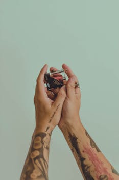 Close-up of tattooed hands holding a colorful object against a soft pastel background.
