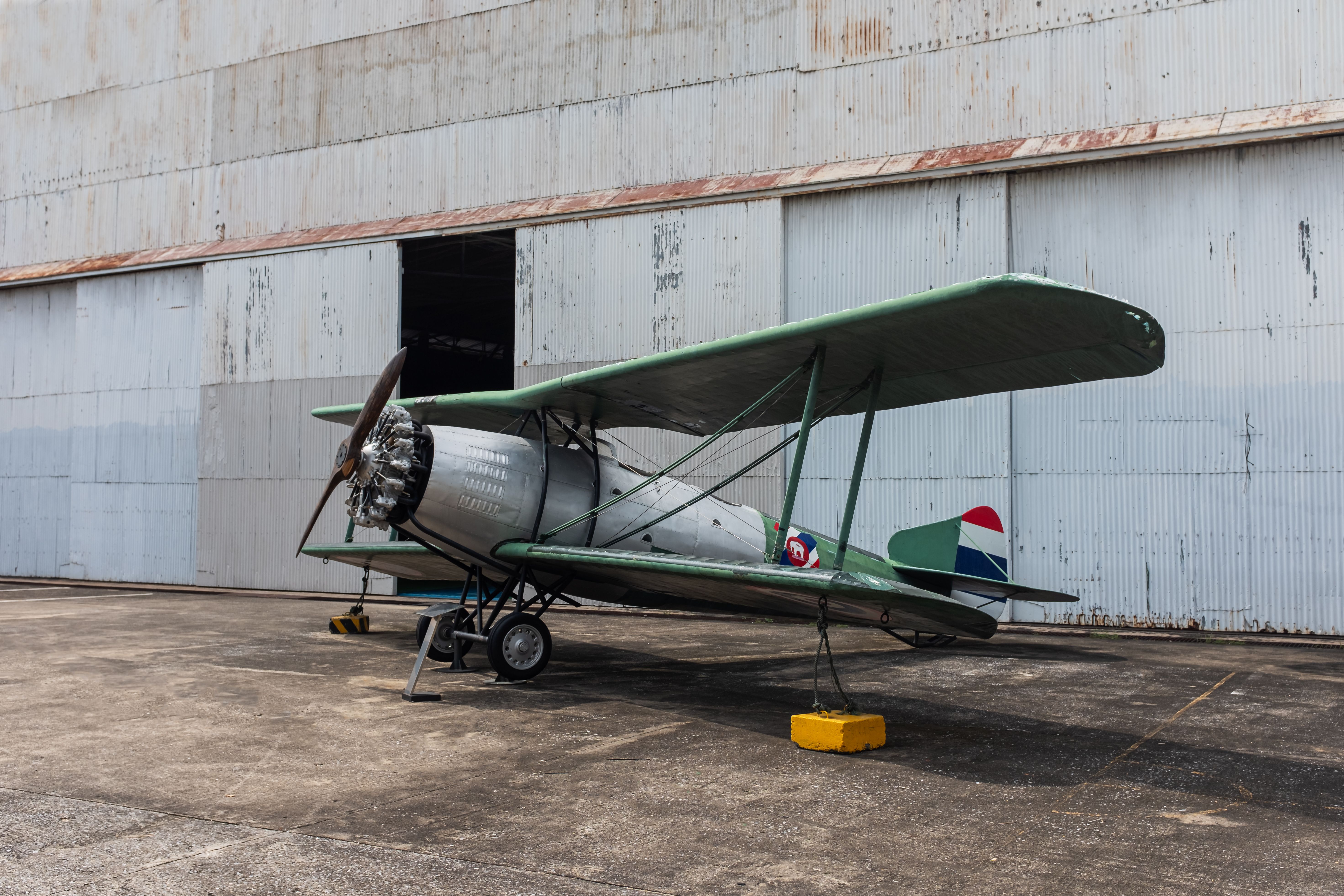 A preserved green biplane with a radial engine and wooden propeller standing on concrete outside a hangar