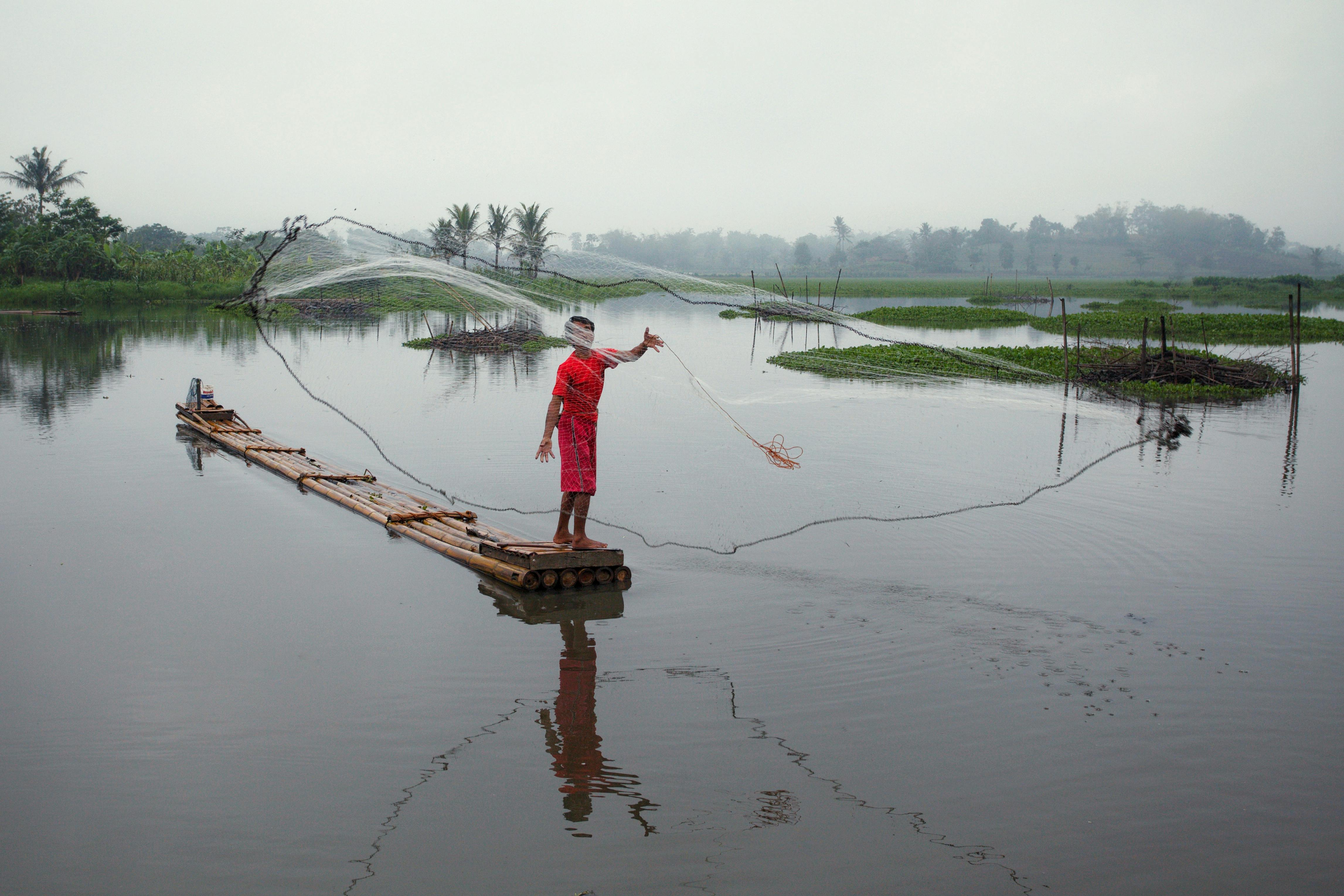 A fisherman casts his net from a bamboo raft on a serene lake surrounded by greenery.