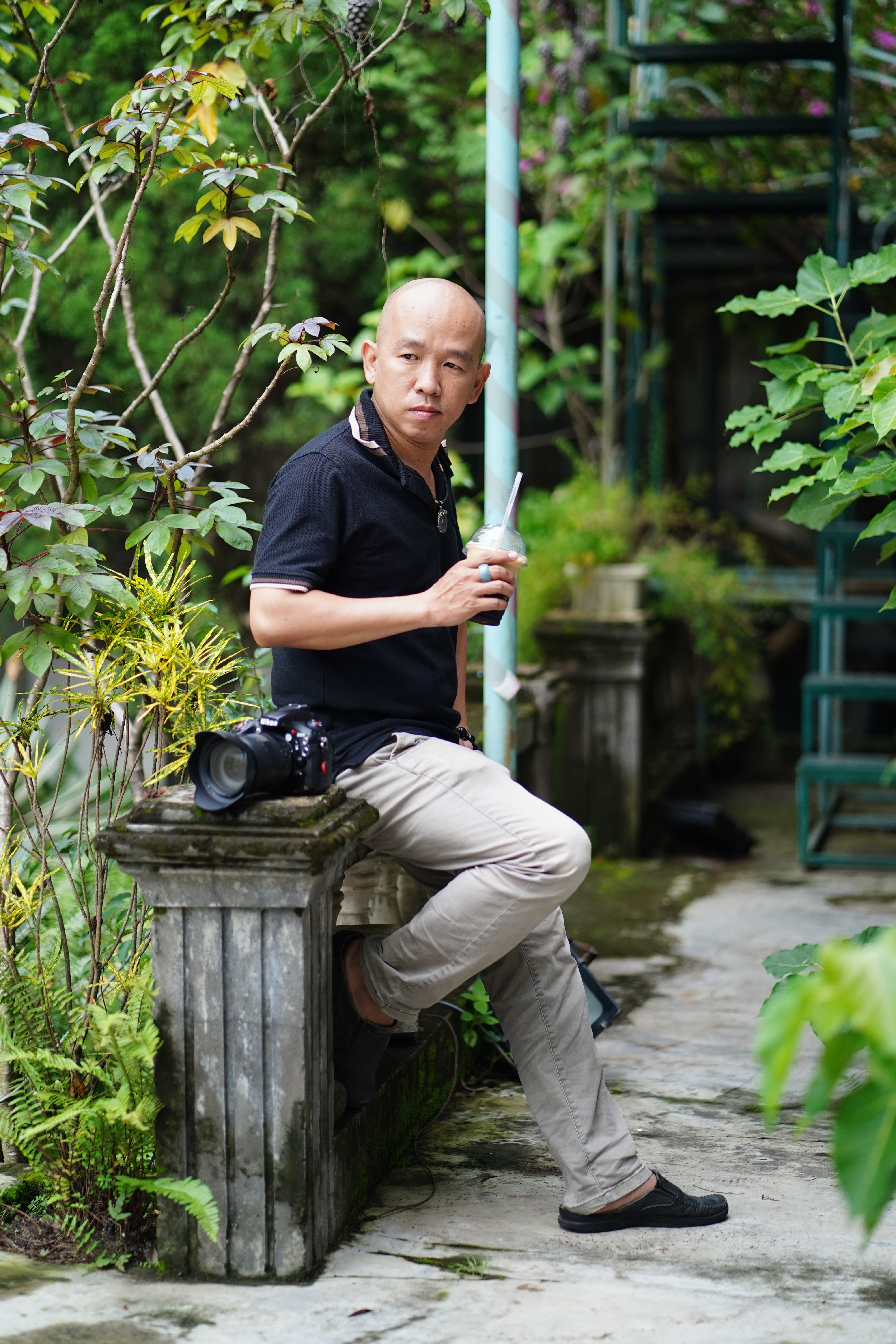 Adult male enjoying a drink in a lush garden setting, with a camera beside him.