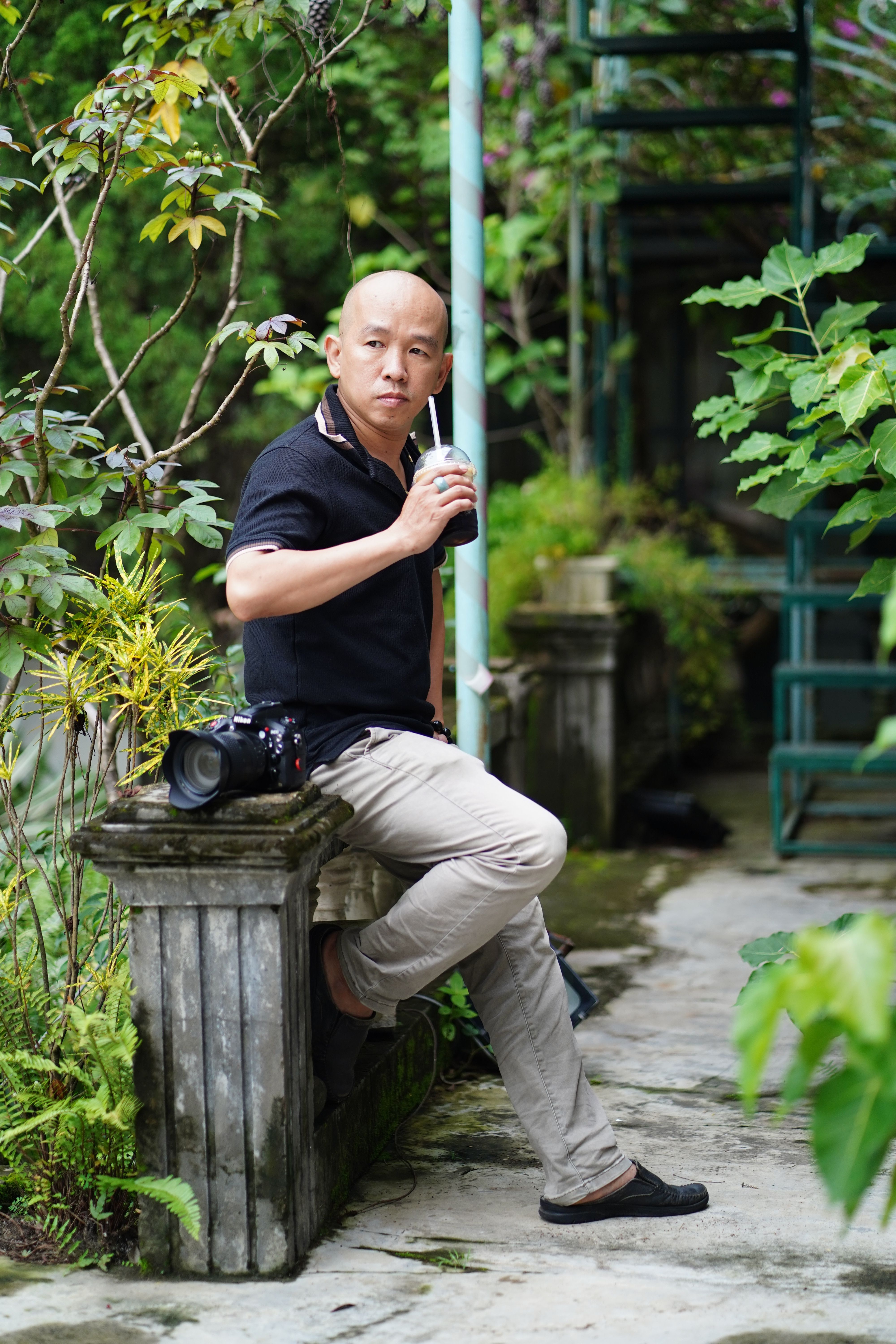 Casual man with a camera takes a break in a green garden, enjoying a drink.