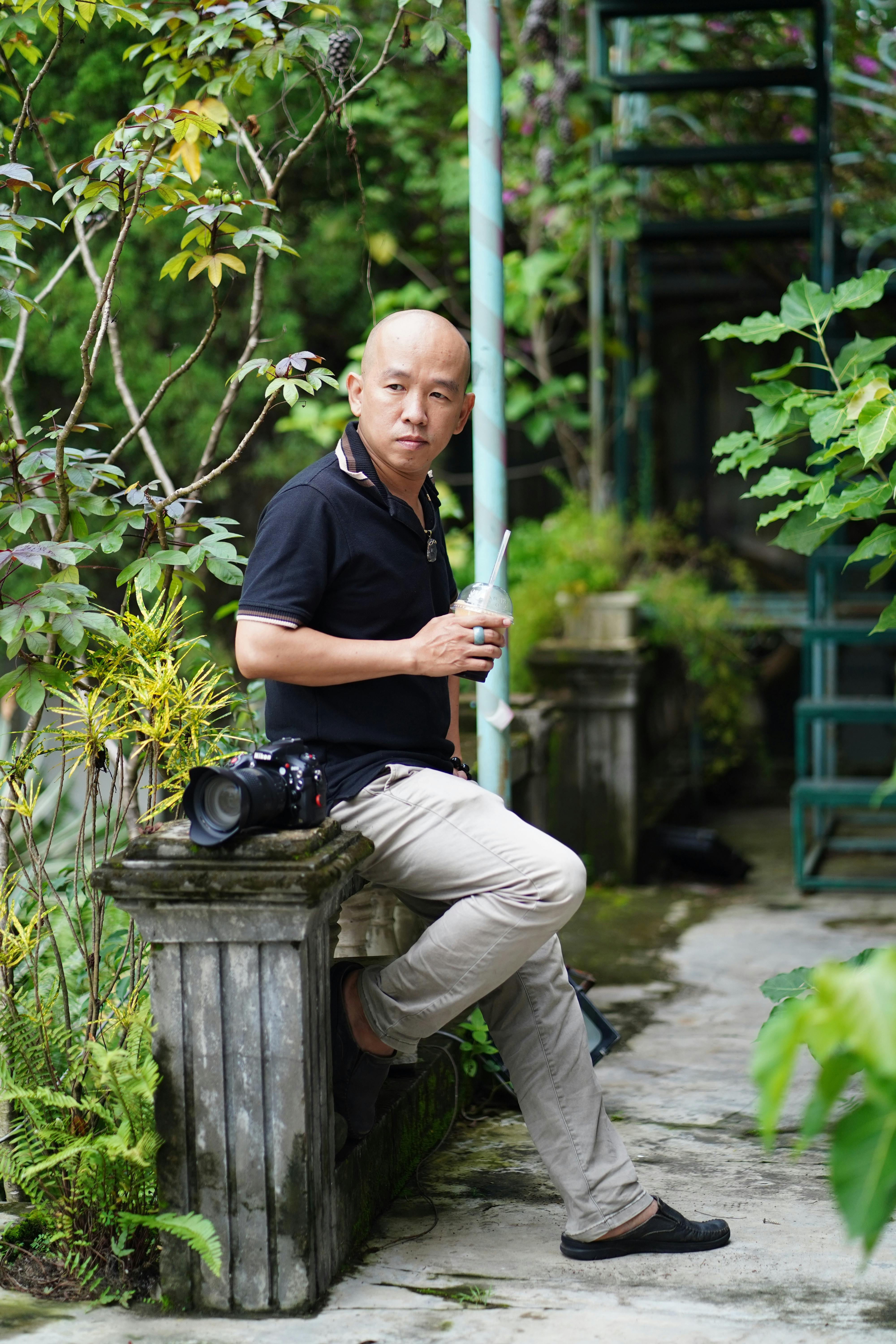 Man sitting in garden with camera, surrounded by greenery. Relaxed and contemplative mood.