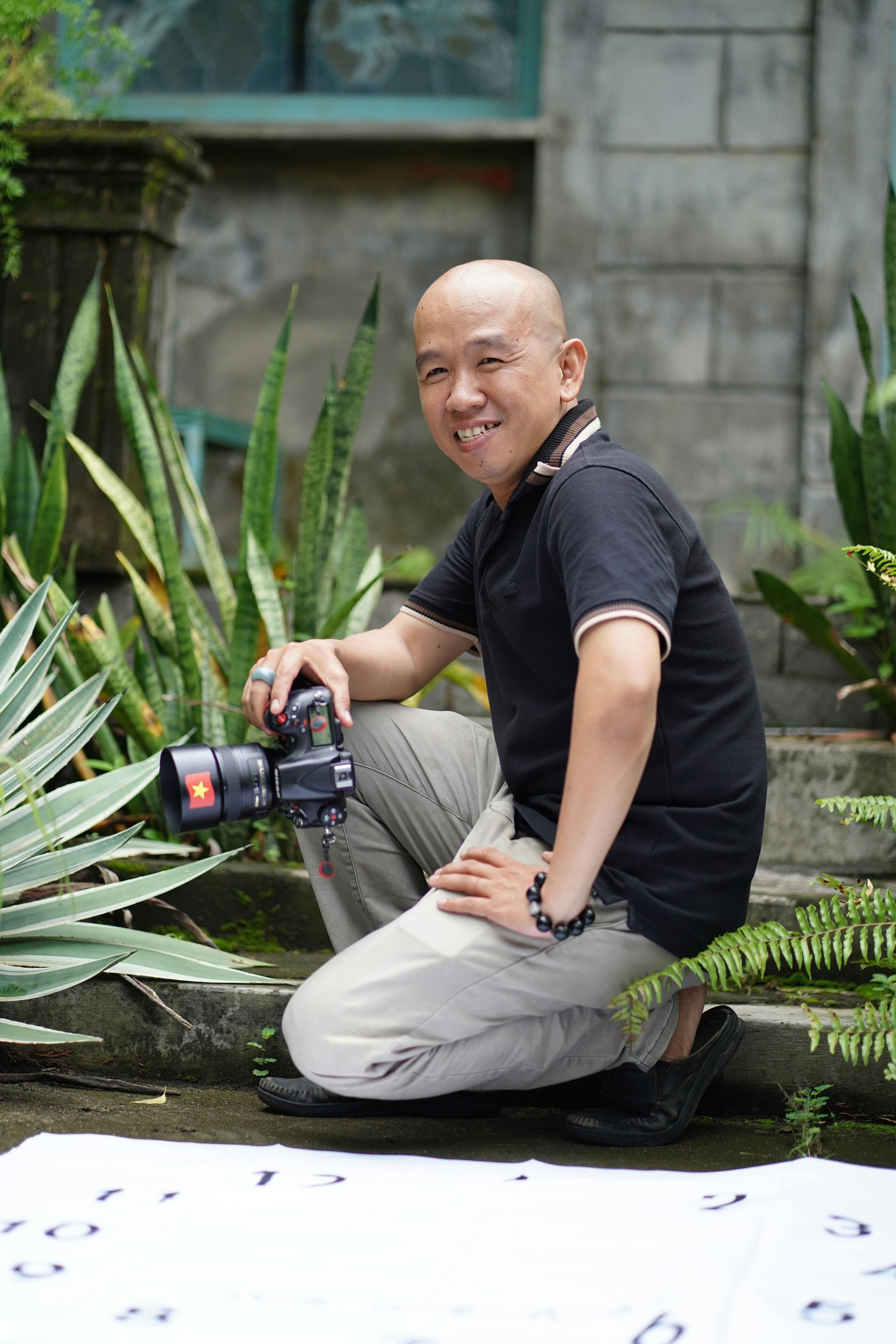 Smiling photographer holding a camera in a lush outdoor garden with greenery.